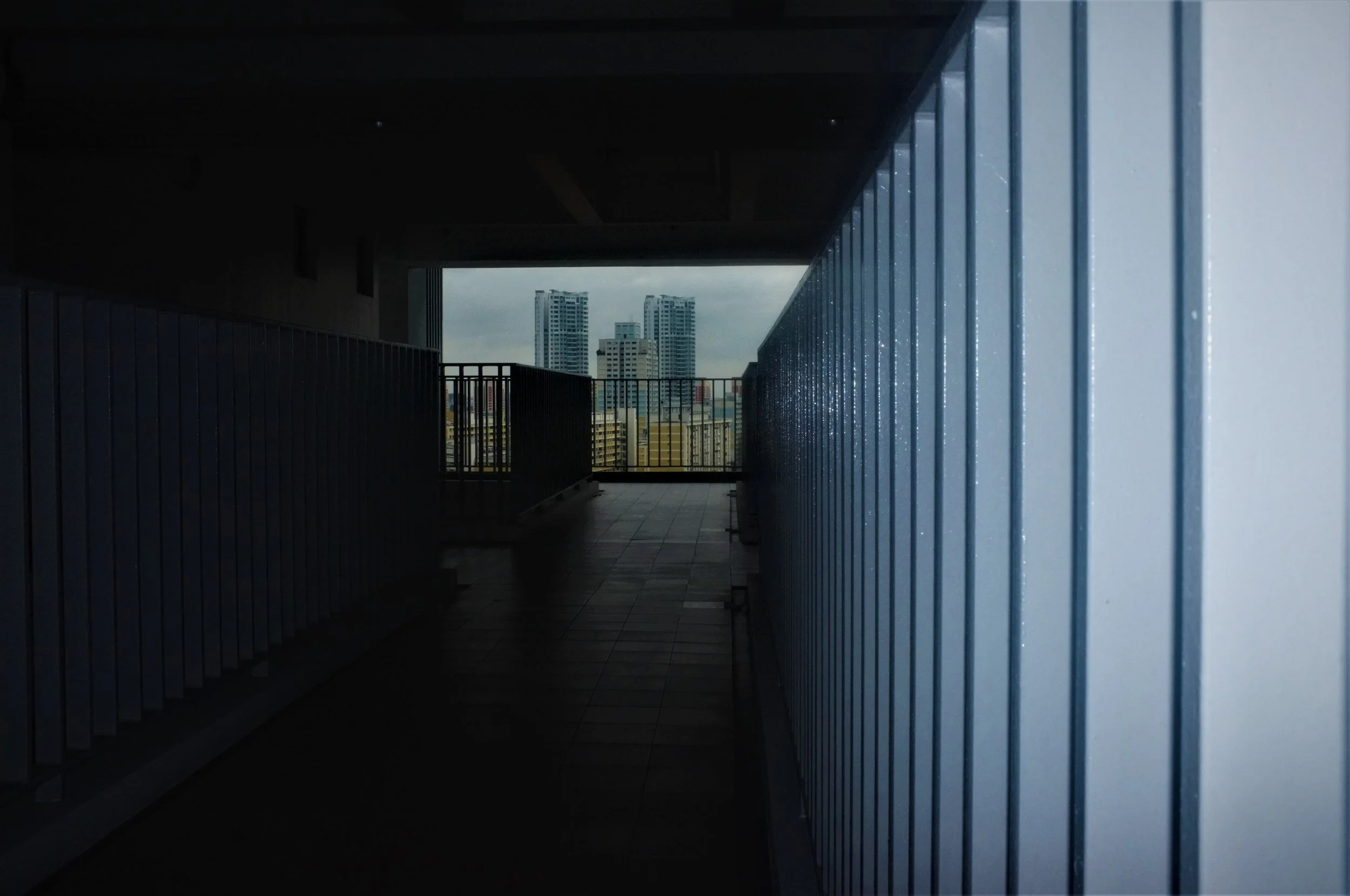 View from dark corridor or balcony with blue metal railing, leading to an open space revealing a cityscape with high-rise buildings under a cloudy sky.