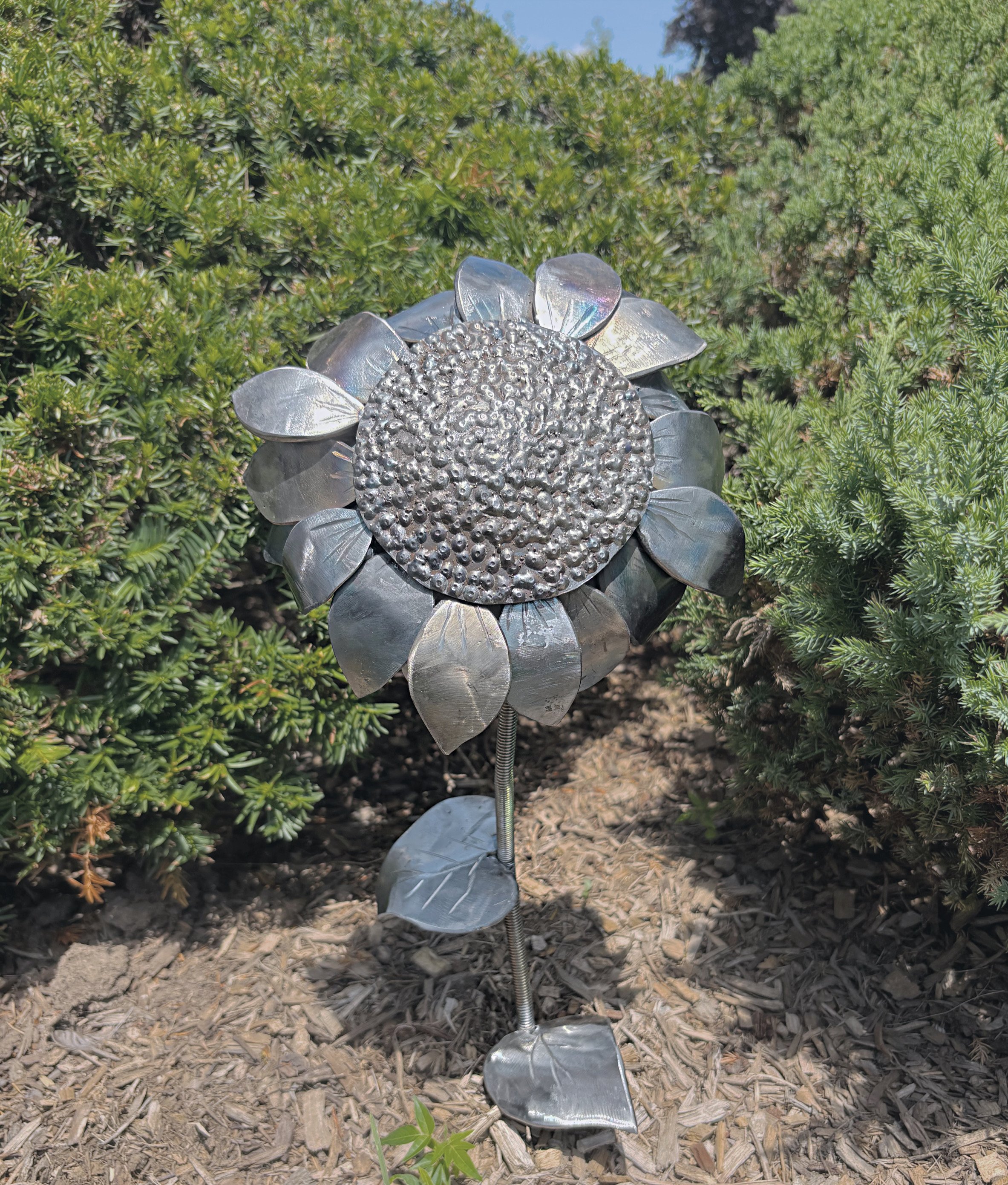 Metal flower sculpture with a textured center and petal-shaped edges placed on a garden bed with mulch, surrounded by green bushes.
