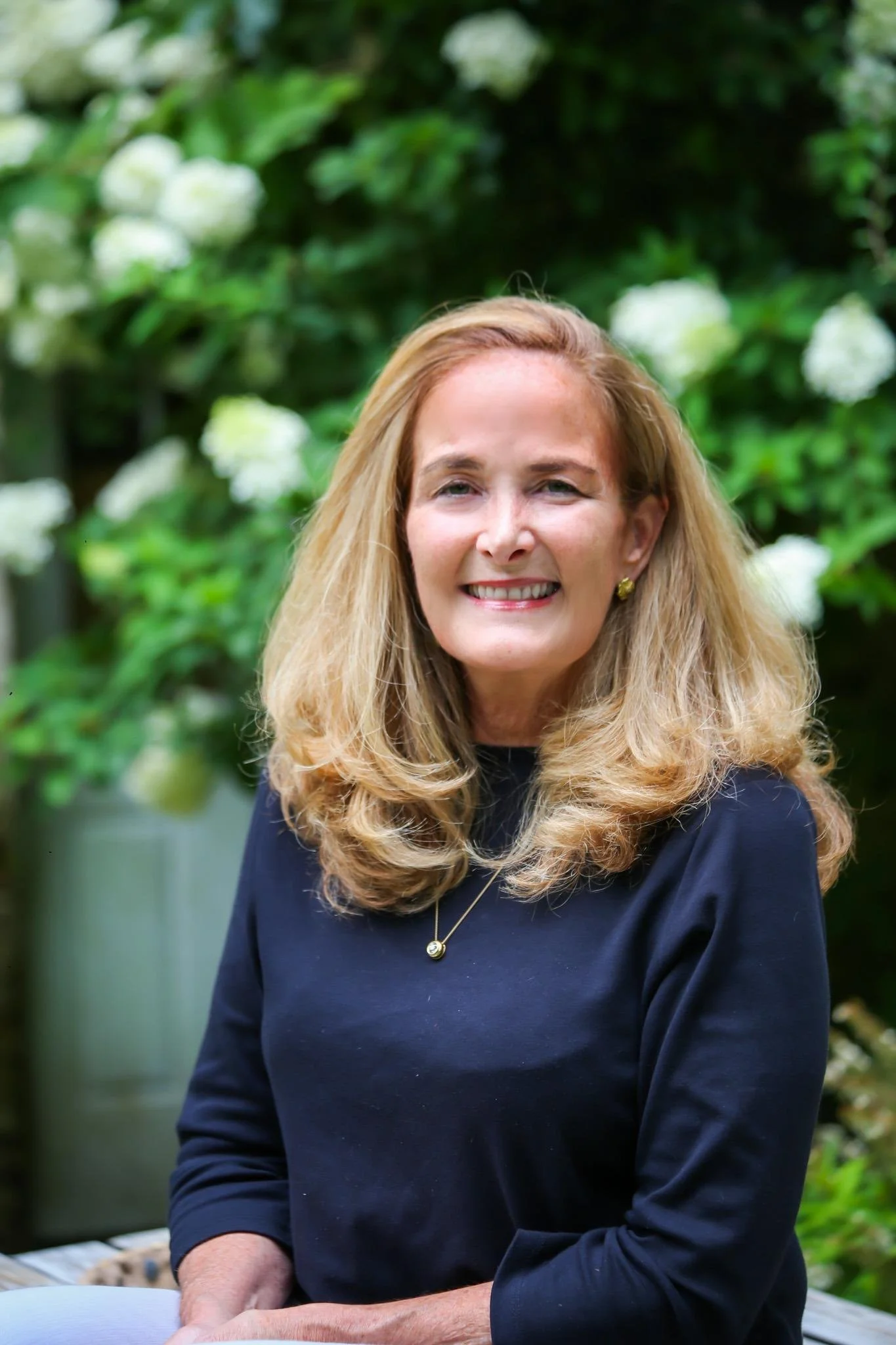 A smiling woman with long blonde hair, wearing gold earrings and a navy blue top, posed outdoors with green leafy plants in the background.