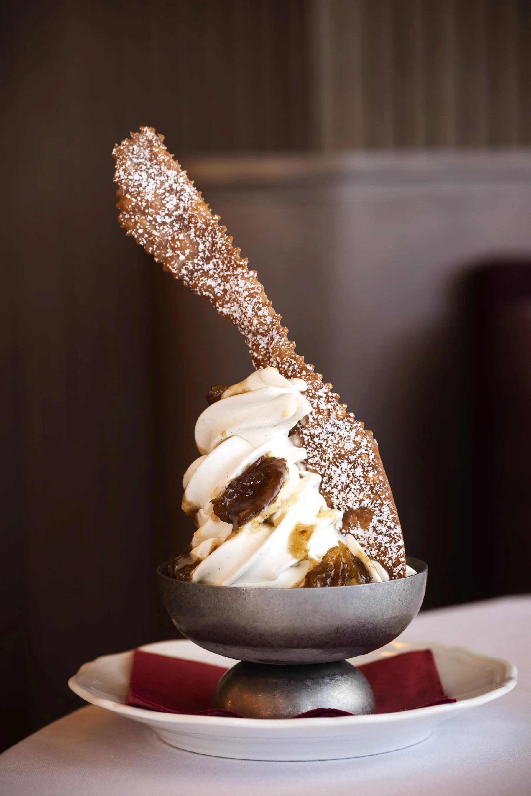 A bowl of ice cream topped with a caramel-coated churro sprinkled with powdered sugar, with caramel sauce drizzled on top, served on a white plate with a red napkin underneath.