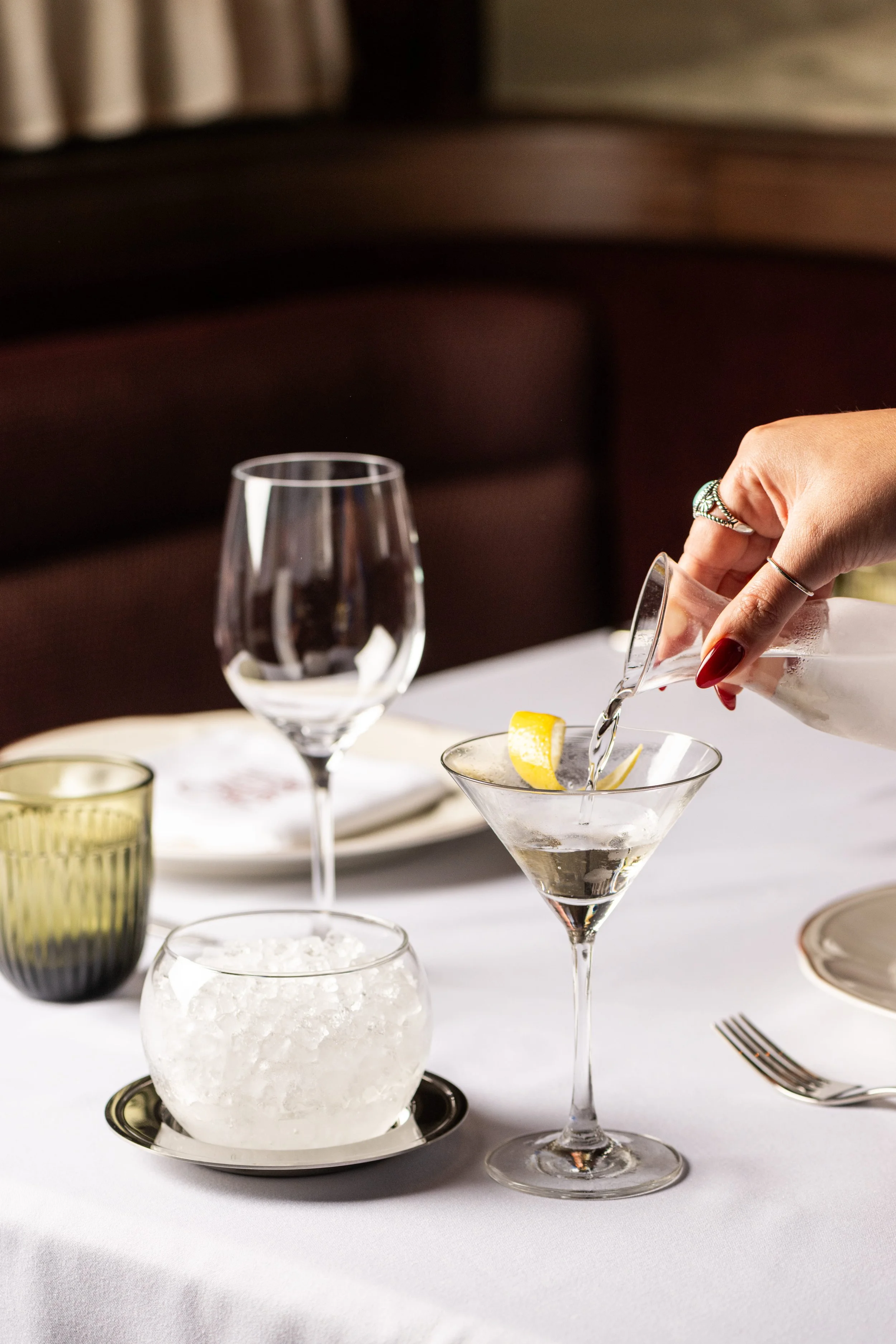 A person's hand with rings pouring a drink into a martini glass with a lemon slice, on a white table with empty glasses, a bowl of ice, and silverware, in a dimly lit setting.