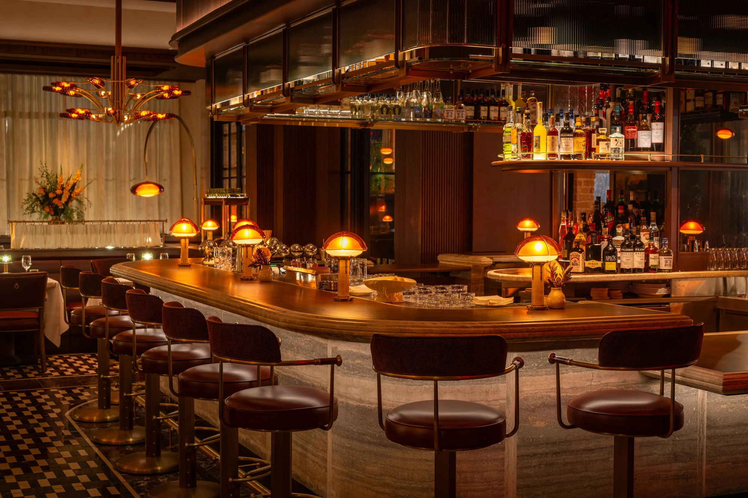 Interior of a bar with a curved wooden counter, bar stools, warm lighting, shelves with liquor bottles, and a decorative chandelier.