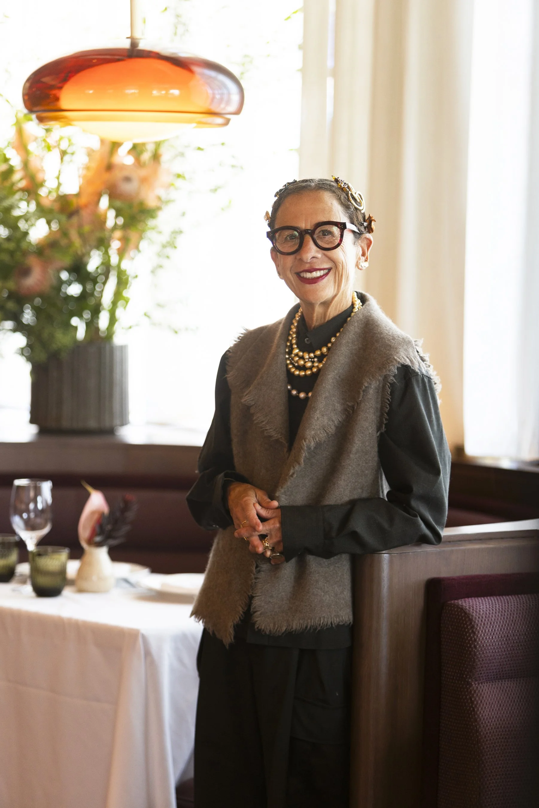 A woman smiling, dressed in black and wearing a brown fur vest, with glasses, jewelry, and hair accessories, standing indoors near a dining table with flowers and tableware.