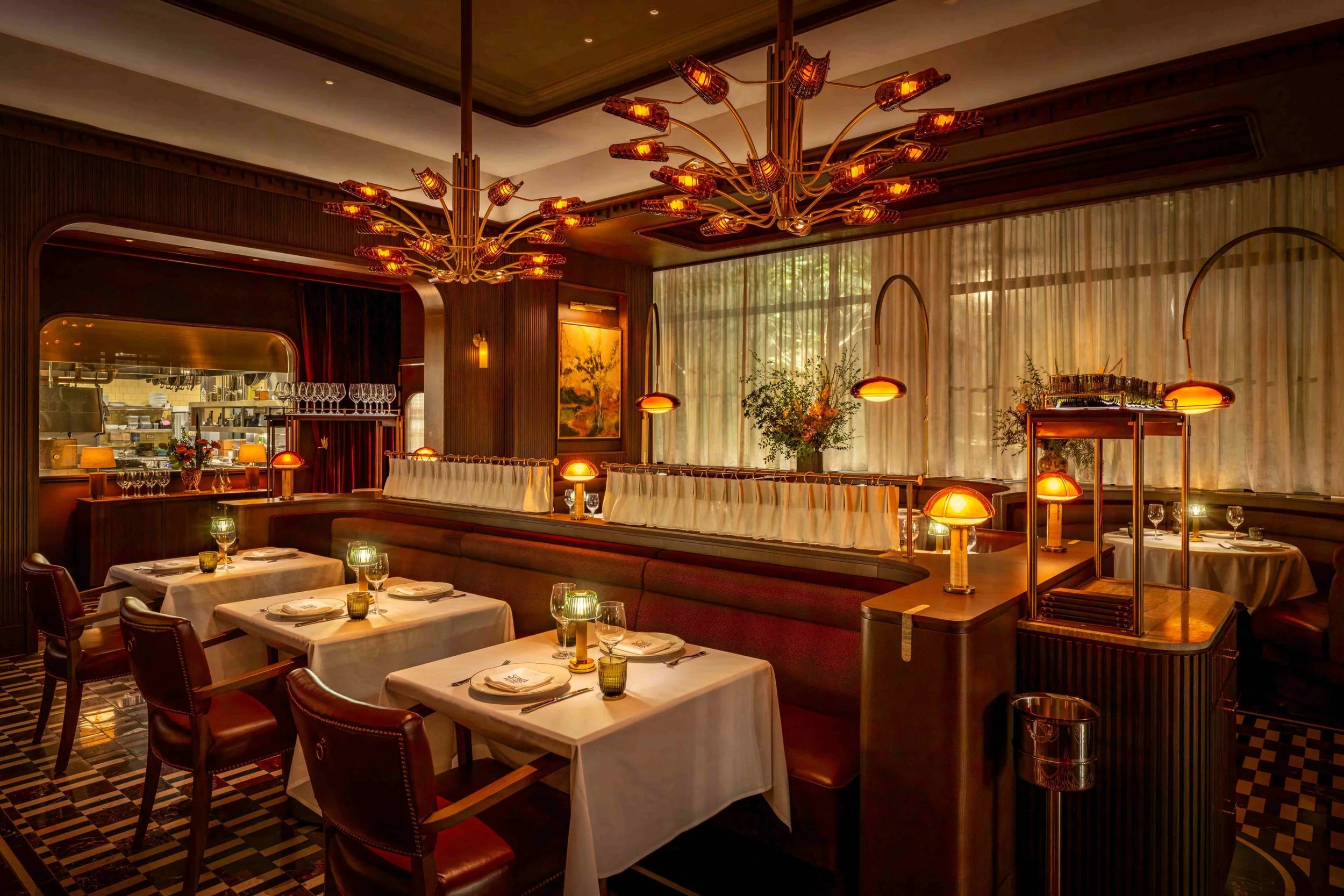 Elegant restaurant interior with warm lighting, decorated with chandeliers, plants, and neatly set tables with white tablecloths, glassware, and candles.