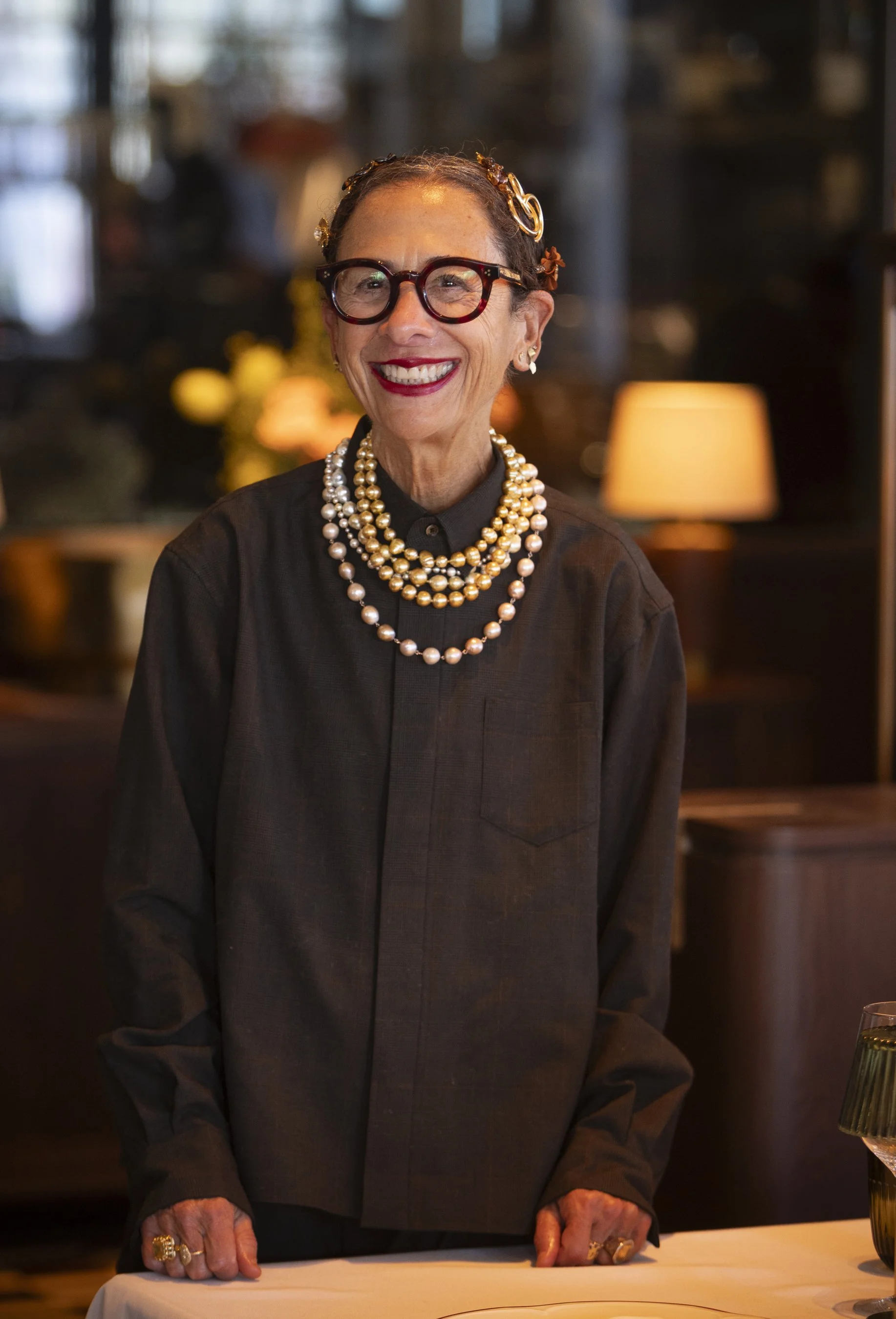 An elderly woman with short gray hair, wearing red glasses, a black shirt, and multiple pearl necklaces, standing behind a table with a white tablecloth and smiling. The background includes warm lighting and blurred decor.