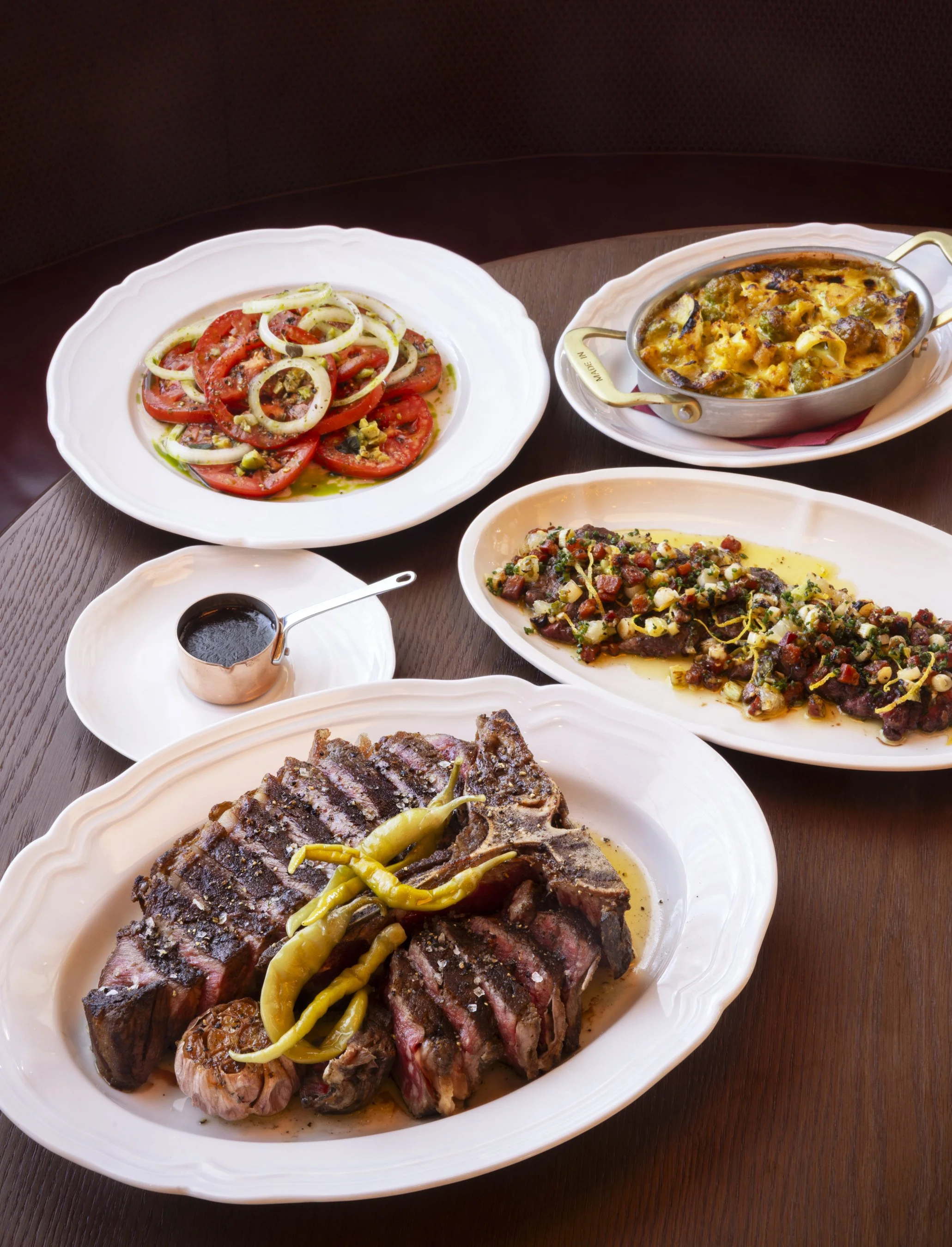 A variety of plates with food on a dark wooden table, including a grilled steak topped with yellow peppers, a dish of baked pasta with cheese and ground meat, a fresh tomato and onion salad, and a small bowl of dark sauce.