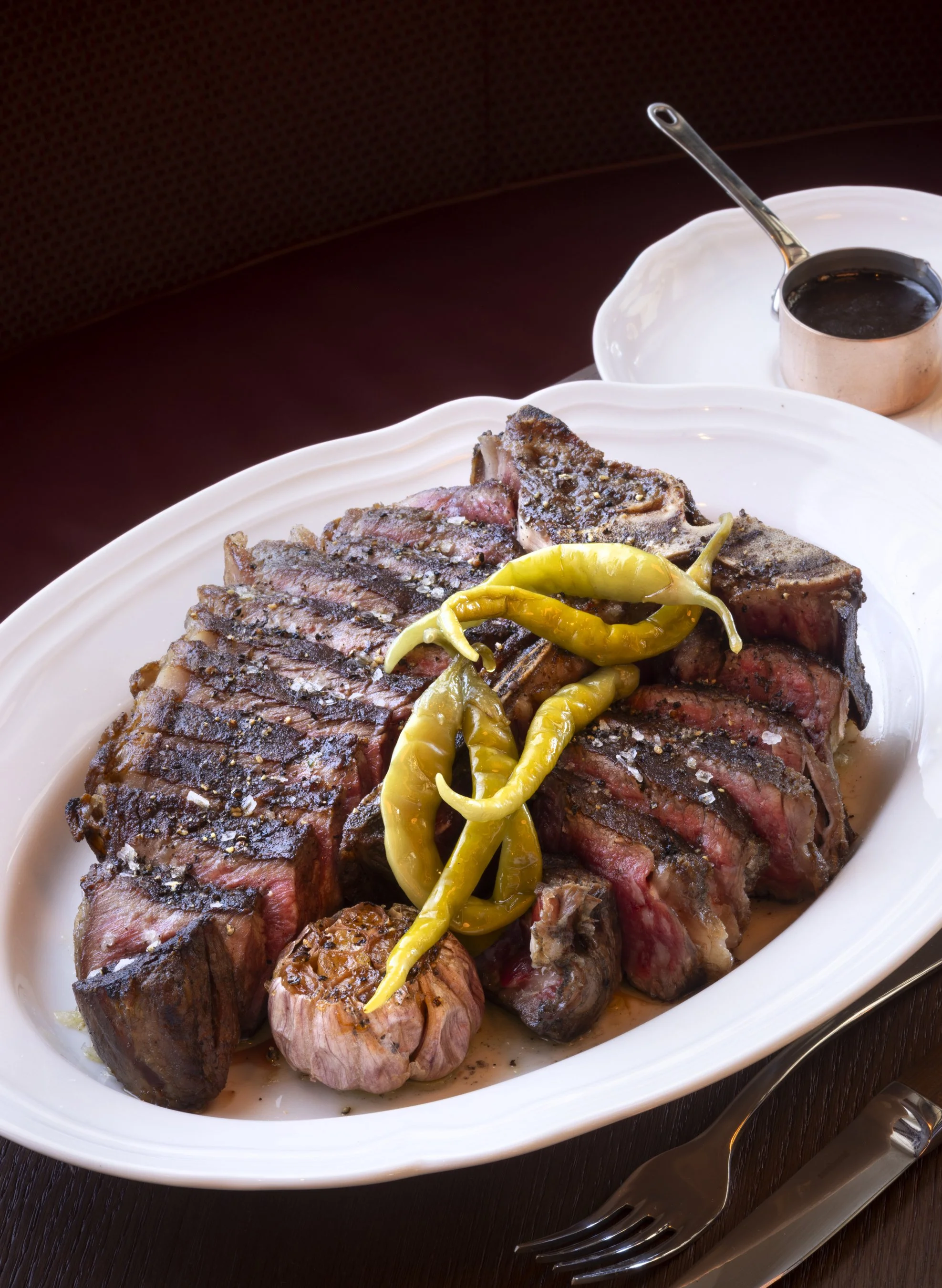 A white oval platter with cooked steak topped with salt and pepper, garnished with green chili peppers, on a dark wooden table beside a fork and knife, with a small copper pot filled with sauce.