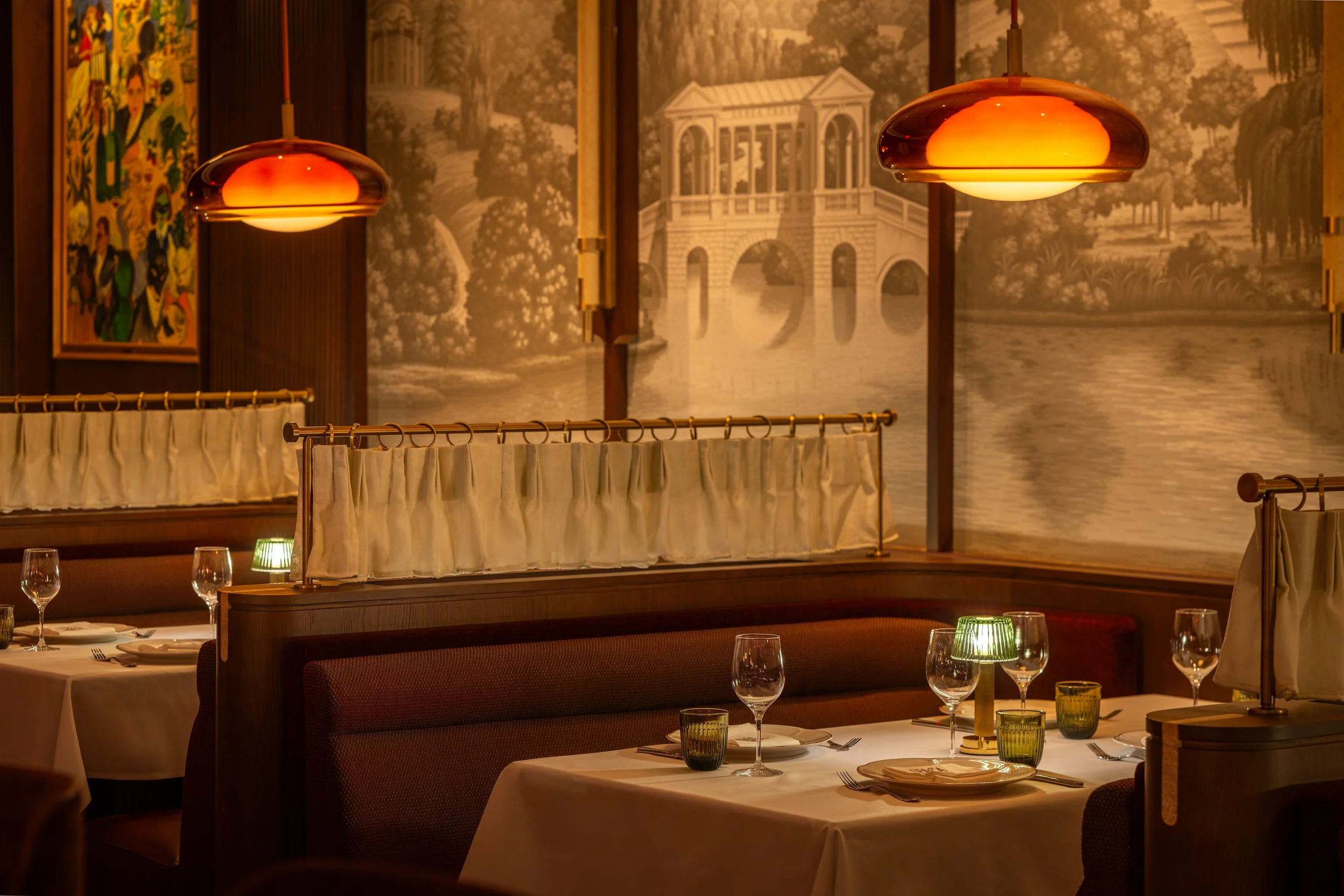 Elegant restaurant interior with warm lighting, vintage landscape mural, and decorated tables set with glasses, plates, and cutlery.