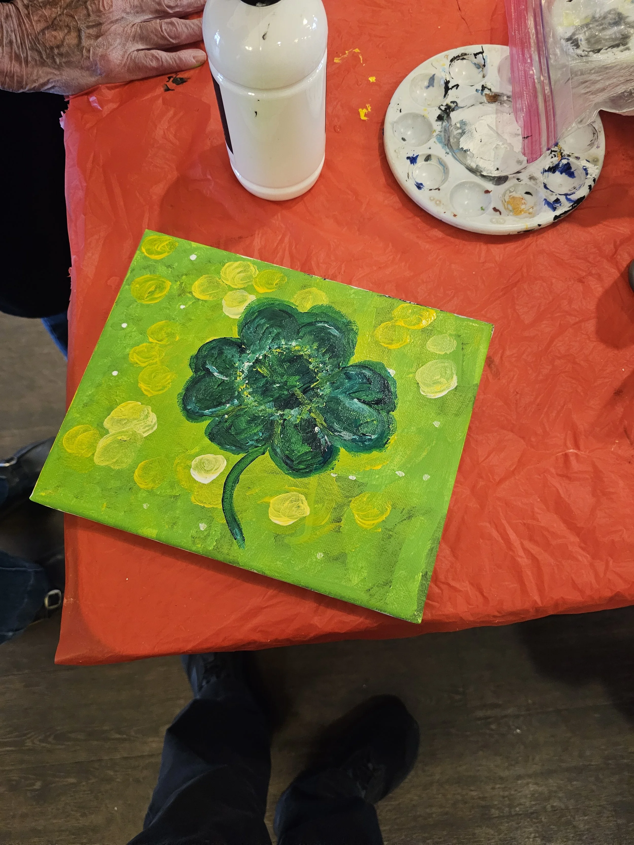 A colorful painting of a green four-leaf clover on a yellow-green background with yellow circles, sitting on a red table. Surrounding the painting are an artist's palette with mixed paints, a bottle of white paint, a small paintbrush, and a plastic b