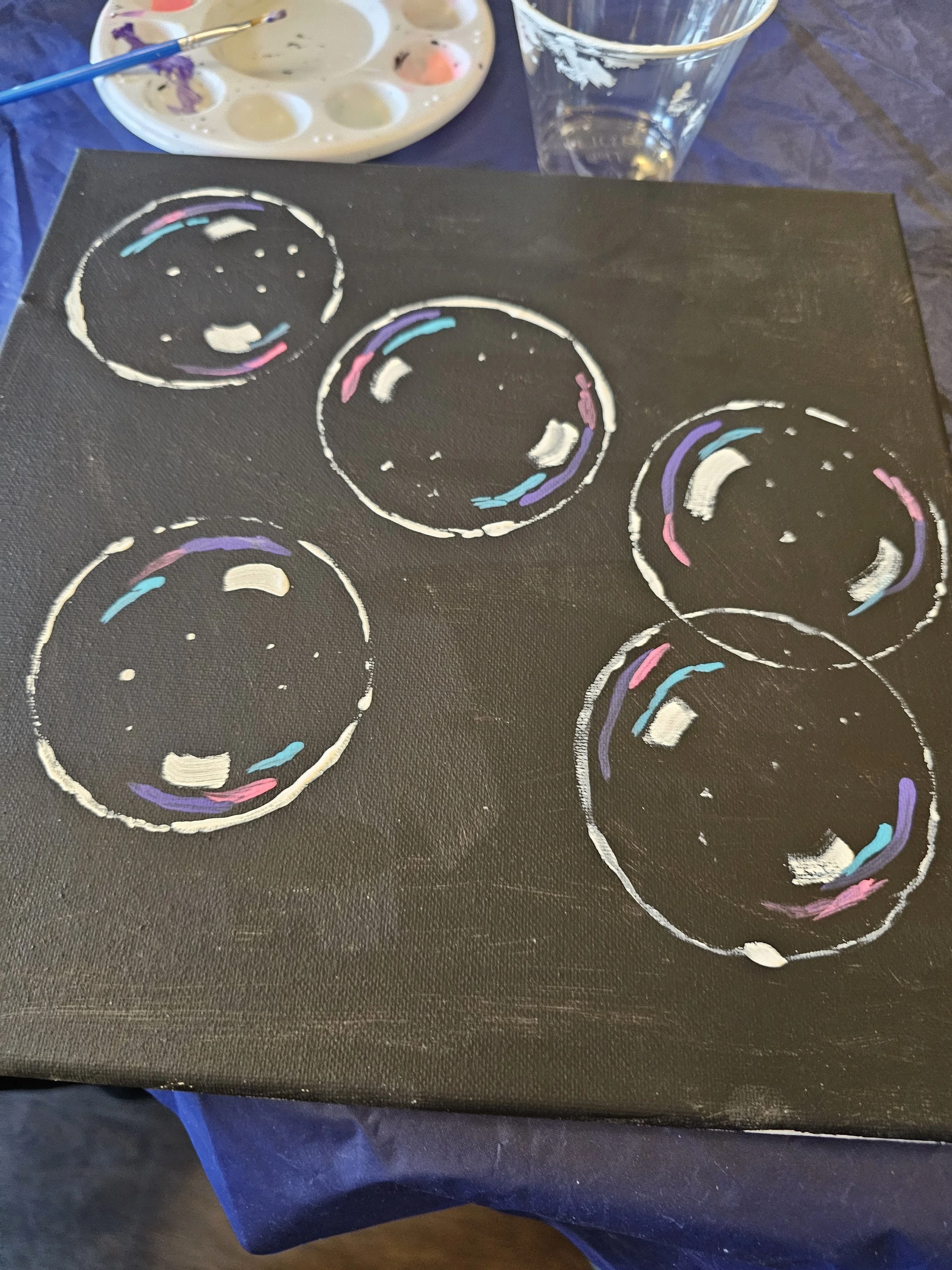 Black canvas with several white circles outlined in white paint, with some colorful accents inside the circles, representing donuts. Art supplies and a glass of water are visible in the background.