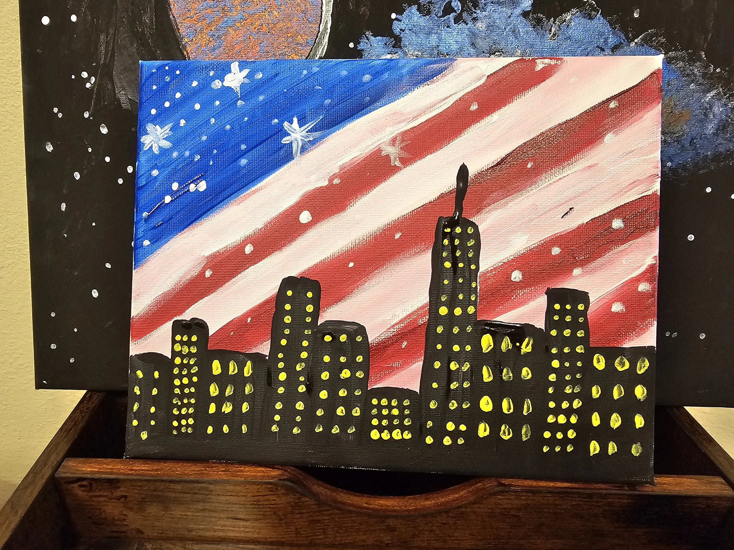 Painting of a city skyline silhouette with bright yellow windows against a background of a starry night sky and an American flag painted with red and white stripes and a blue field with white stars.