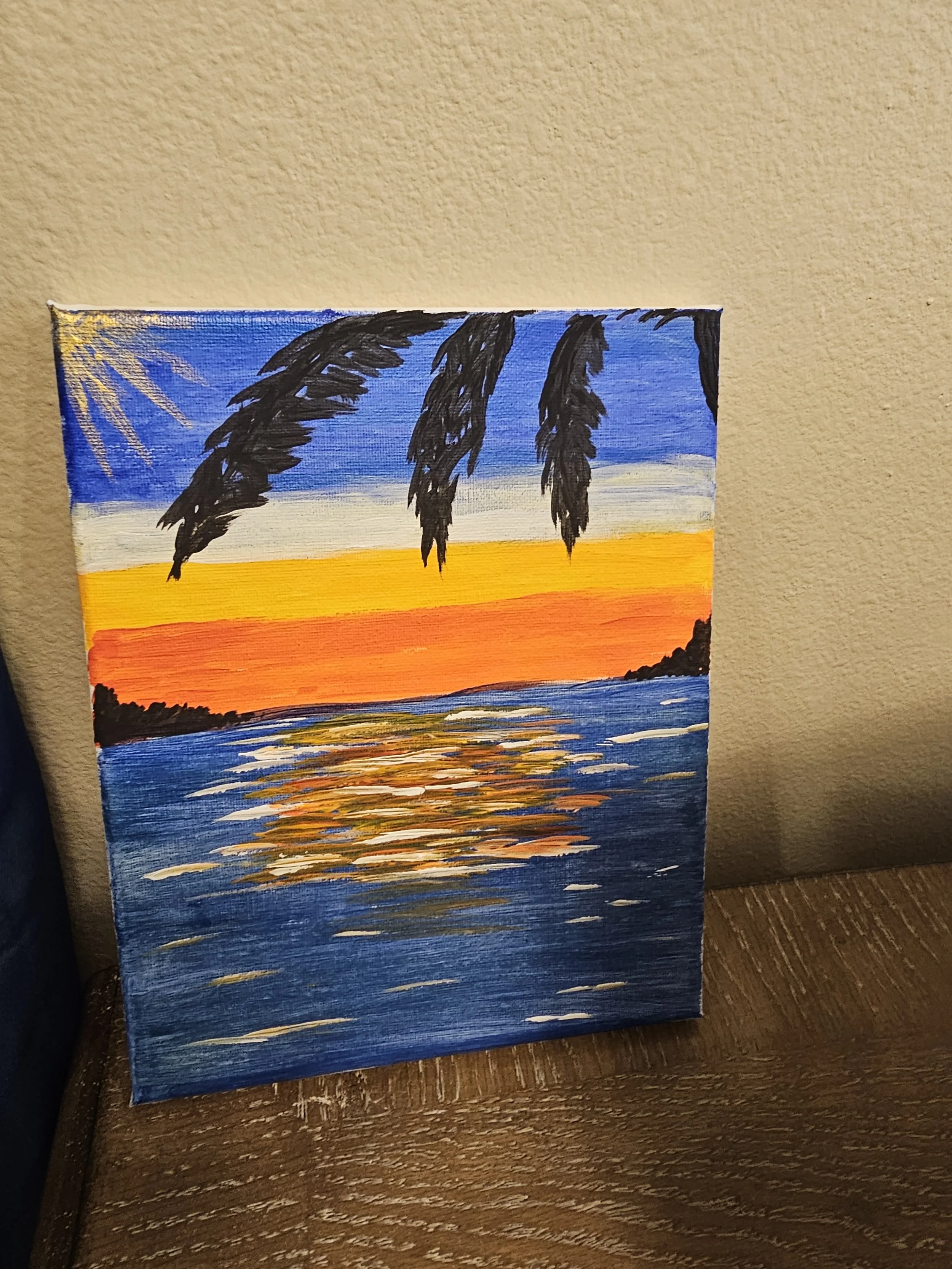 A painting depicts a beach scene at sunset with a bright orange and yellow sky, a silhouette of trees along the shoreline, and water reflecting the colors of the sky.