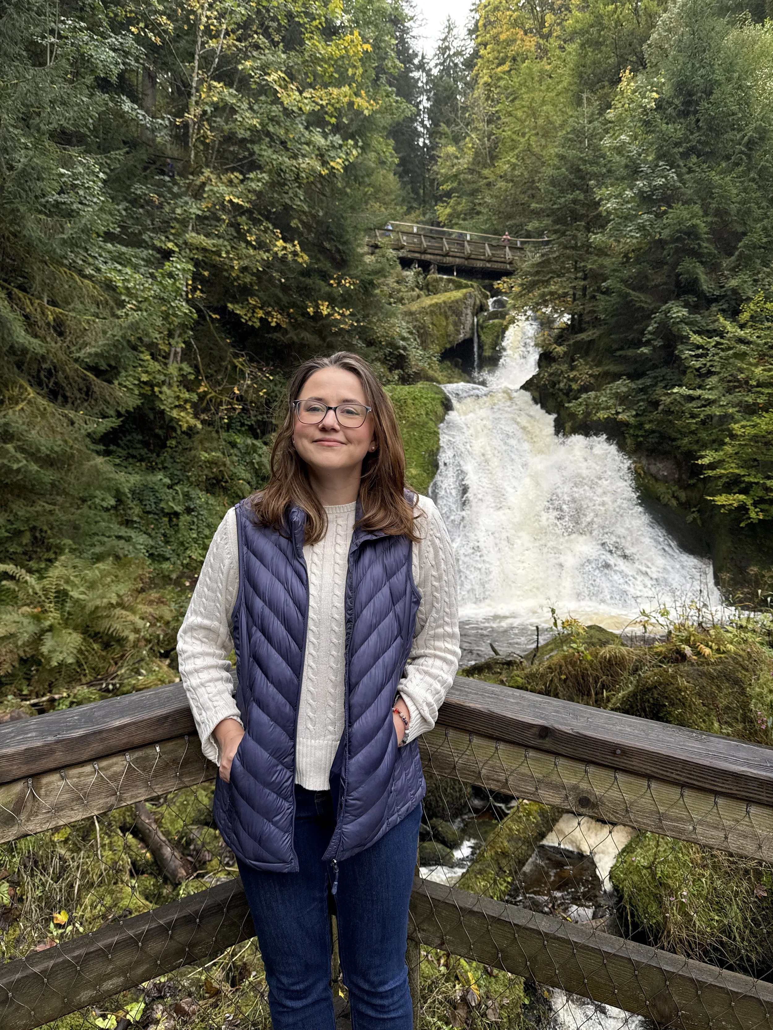 A woman standing on a wooden bridge in front of a waterfall surrounded by lush green trees and foliage.