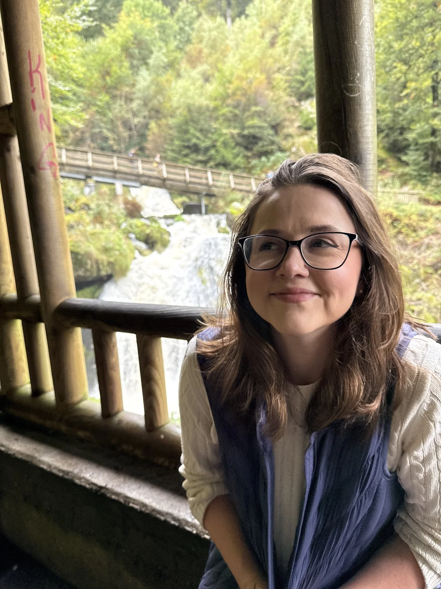 A young woman wearing glasses, a white shirt, and a dark vest, smiling while sitting inside a wooden structure near a waterfall and lush green trees.