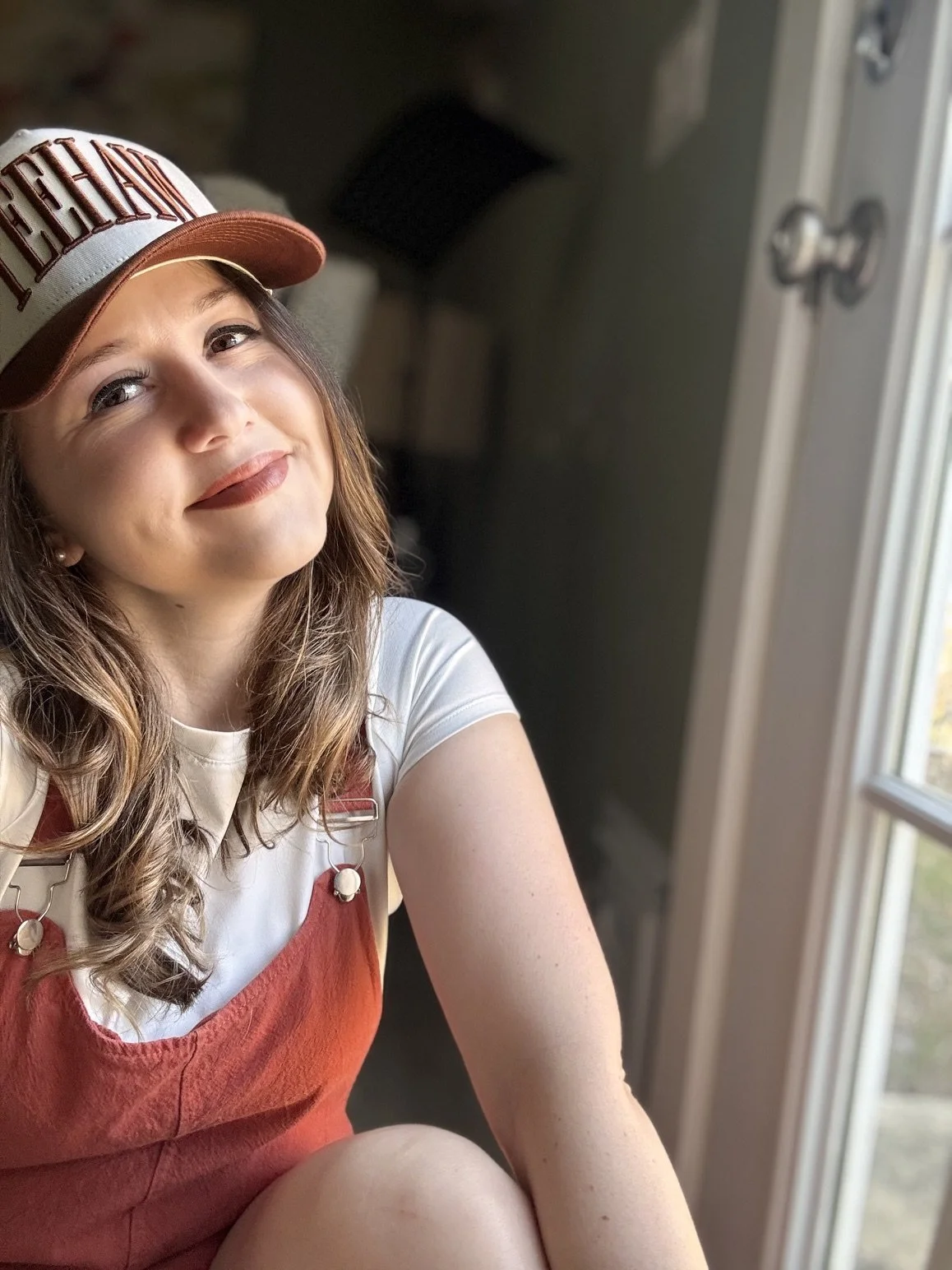 A young woman with wavy brown hair wearing a baseball cap, white shirt, and red apron, sitting near a window with natural light and smiling.