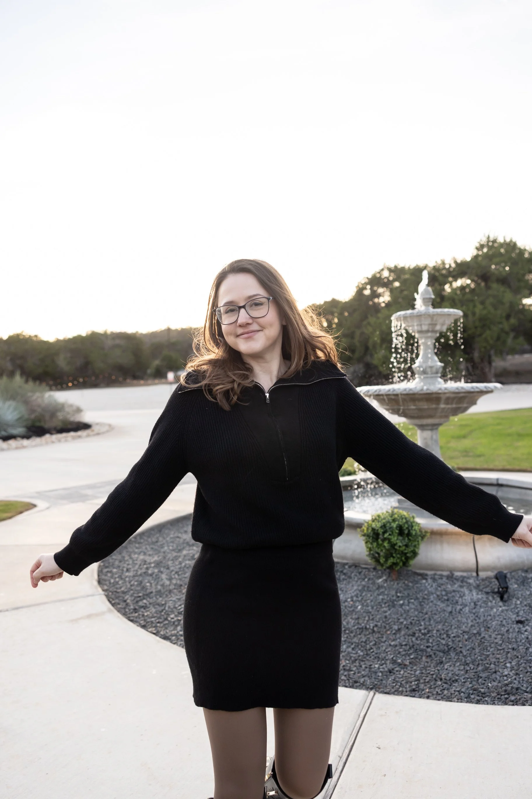 A young woman with glasses and wavy brown hair standing outdoors near a fountain, smiling with her arms outstretched, wearing a black dress and beige tights.
