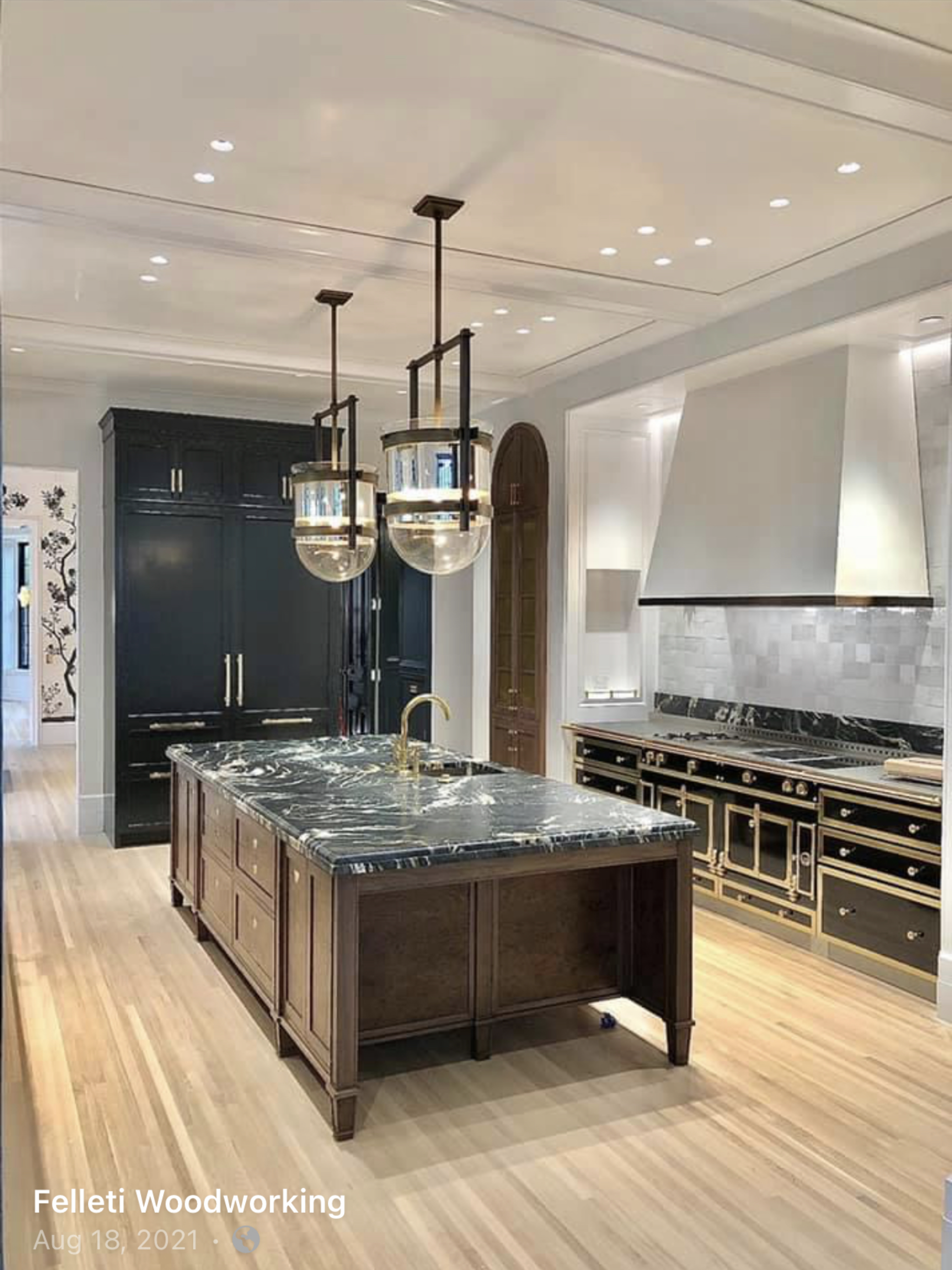 A spacious kitchen with a central island featuring a black marble countertop and a brass faucet. The kitchen has dark cabinetry, stainless steel appliances, a marble backsplash, and a hardwood floor. Ceiling lights are visible, and modern light fixtures hang over the island.
