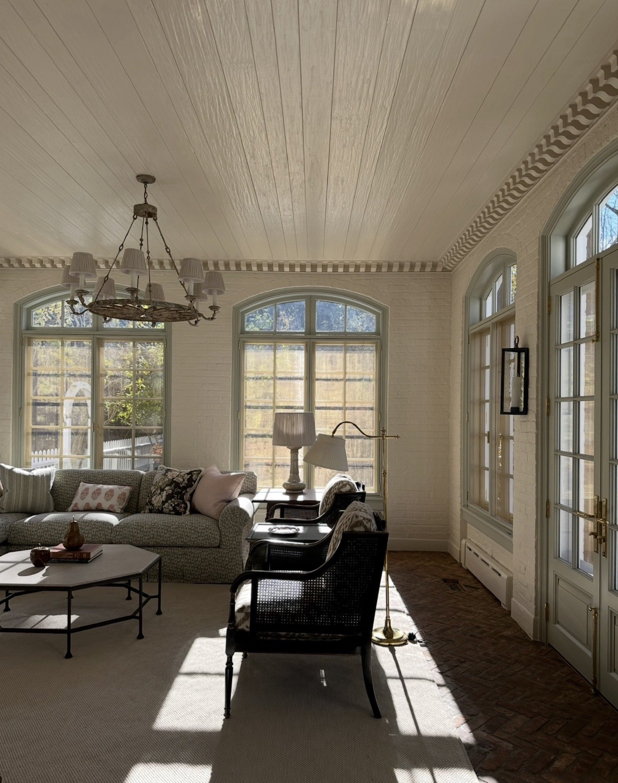 Sunlit living room with large windows, white brick walls, a chandelier, a sofa with cushions, lamps, and chairs on a beige rug.