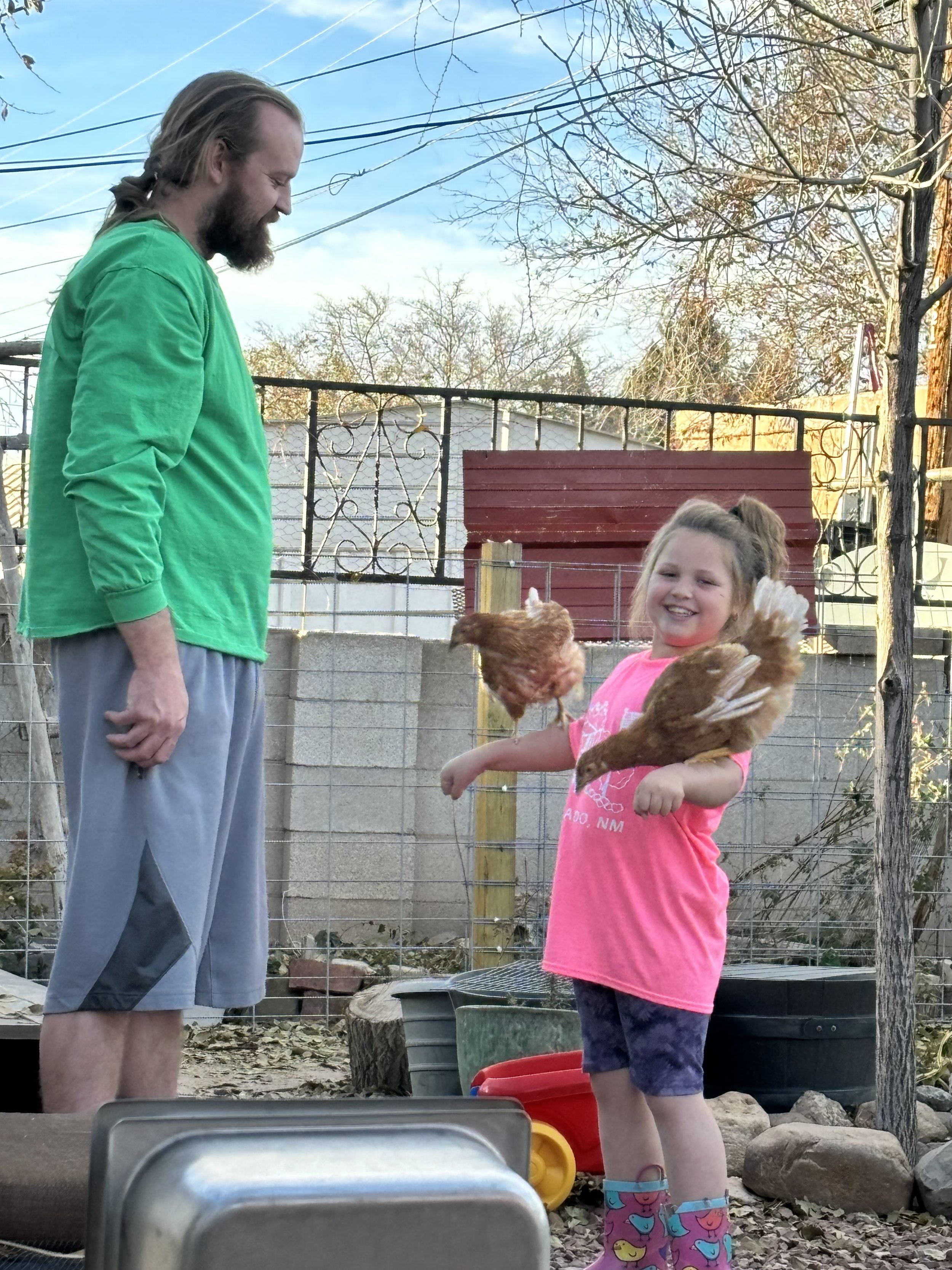 A man and a young girl outside in a backyard, holding chickens. The man is wearing a green shirt and gray shorts, smiling while looking down at the girl. The girl is wearing a pink shirt, patterned rain boots, and is holding chickens, smiling happily.
