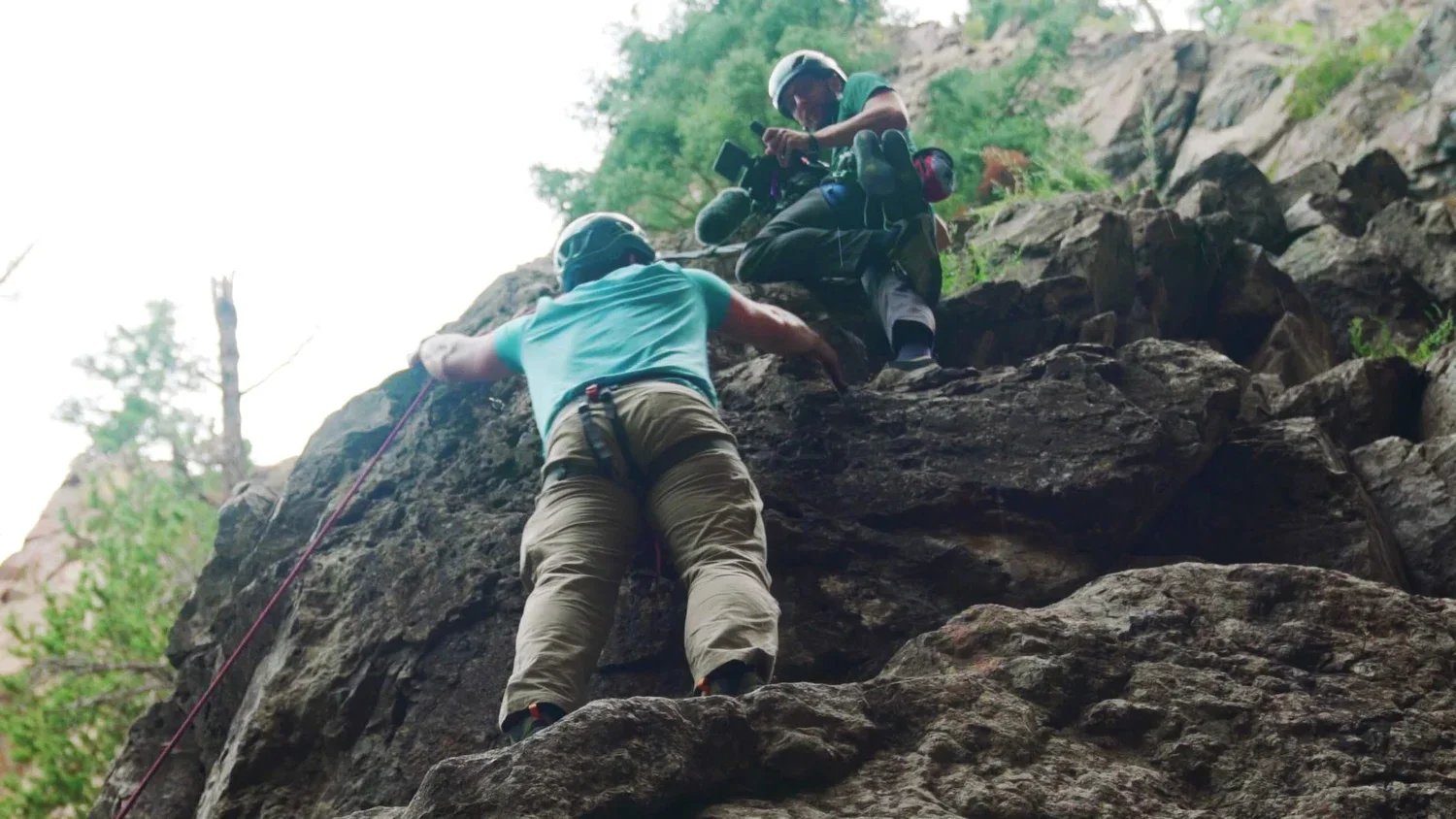 Two rock climbers wearing helmets and harnesses ascending a rocky cliff, one helping the other while holding onto the rock face.
