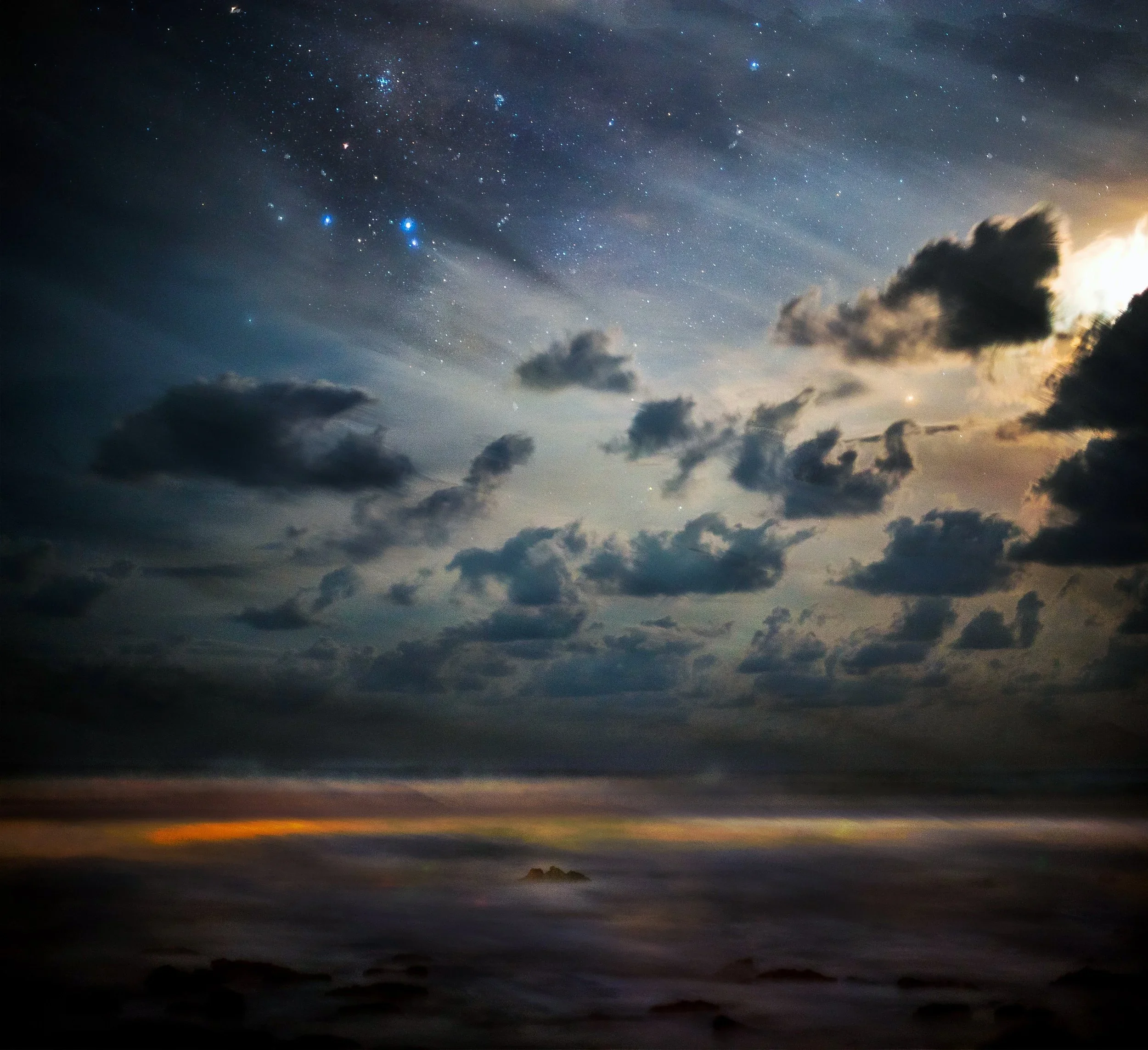 Night sky view over the ocean with stars, clouds, and a faint horizon line.