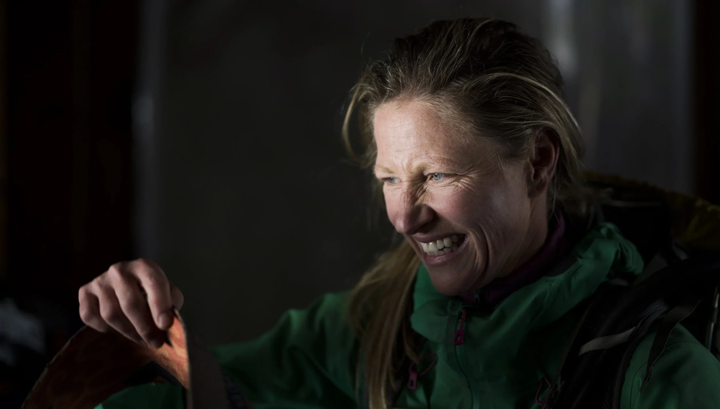 A woman with blonde hair in a ponytail, wearing a green jacket, smiling widely and holding a piece of food in her hand.