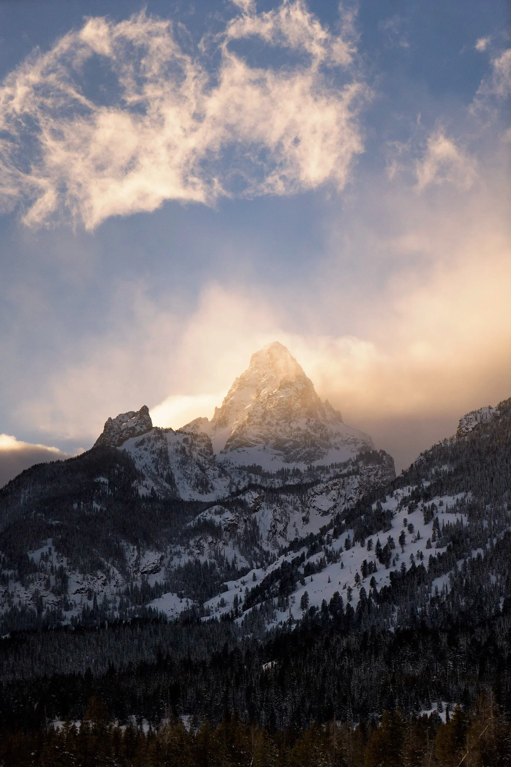 Snow-capped mountain peak with partly cloudy sky and a golden glow from the setting sun, surrounded by forested hills.