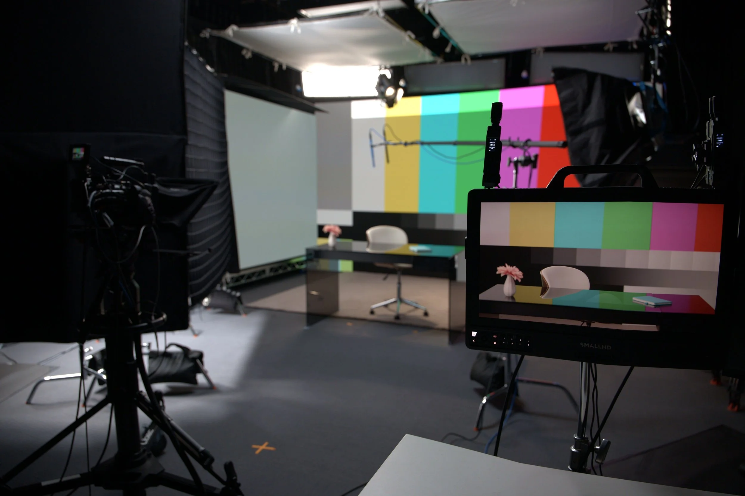 A television studio setup with professional lighting, cameras, and a large screen displaying a test pattern with multicolored bars, a table with chairs, and equipment for filming or broadcasting.