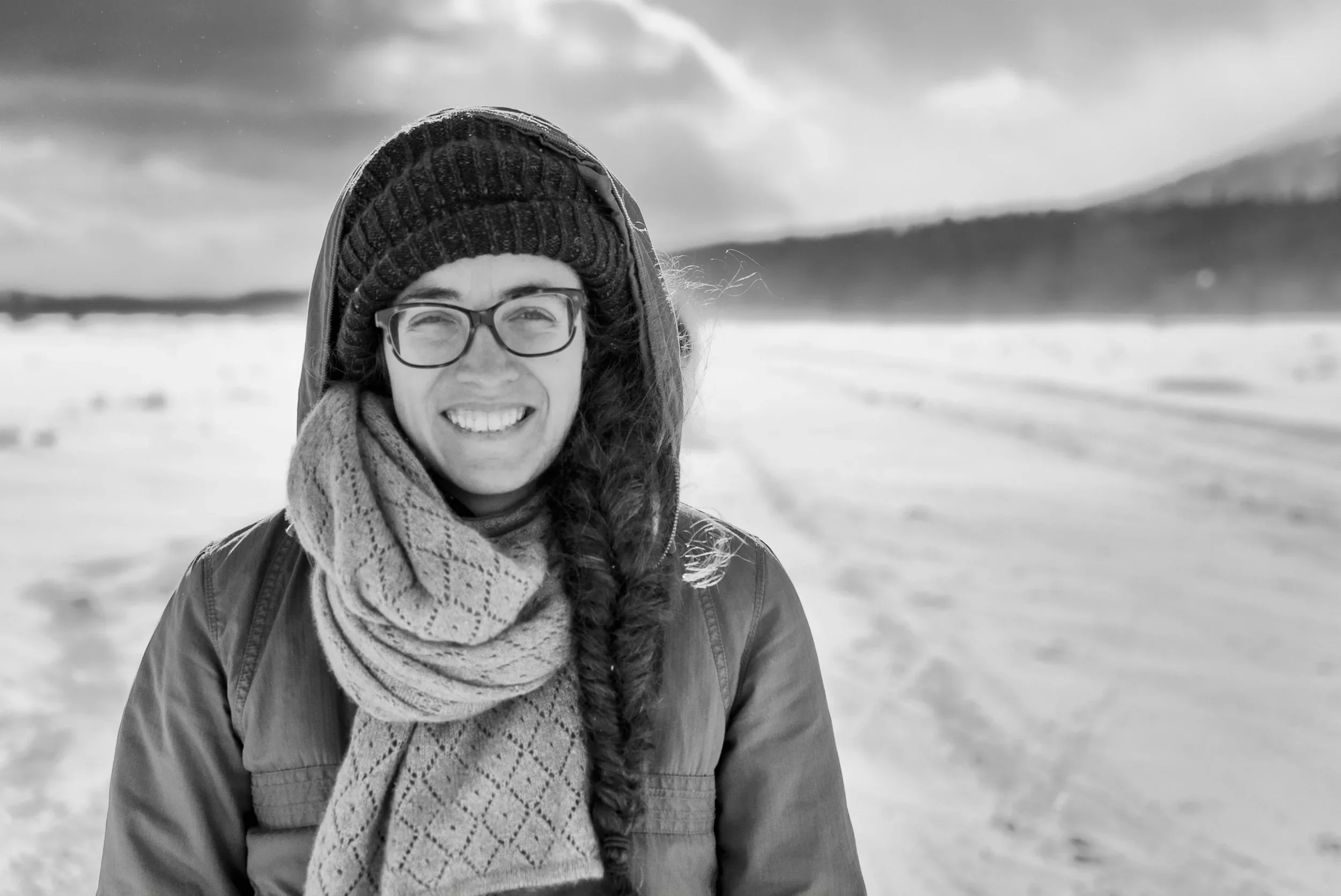 Smiling woman wearing a plaid winter coat, knitted beanie, glasses, and scarf outdoors in a snow-covered landscape with mountains in the background.