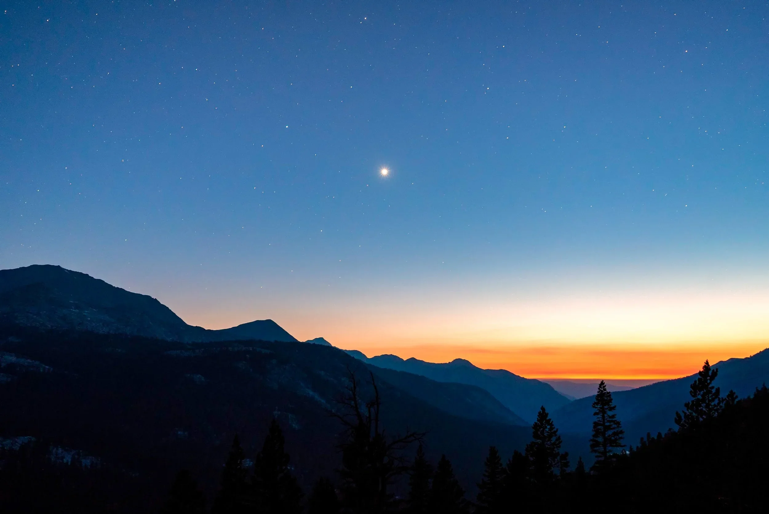 A scenic landscape of mountain ranges during sunset with a clear sky filled with stars and a bright planet or star near the center.