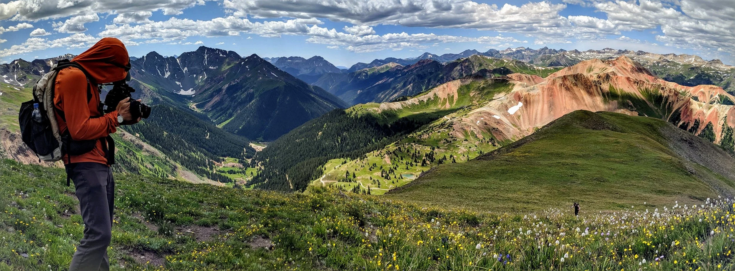 Person wearing orange jacket and backpack taking a photo of colorful mountain landscape with green valleys and wildflowers.