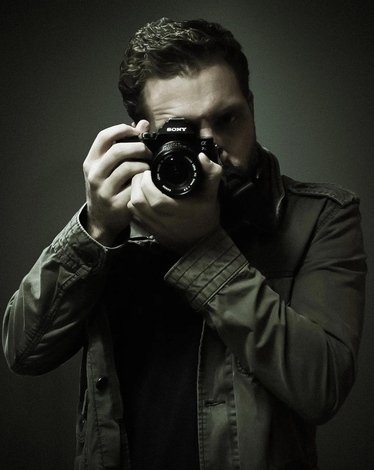 Person taking a self-portrait photo using a Sony camera, wearing a dark jacket, with dark background.