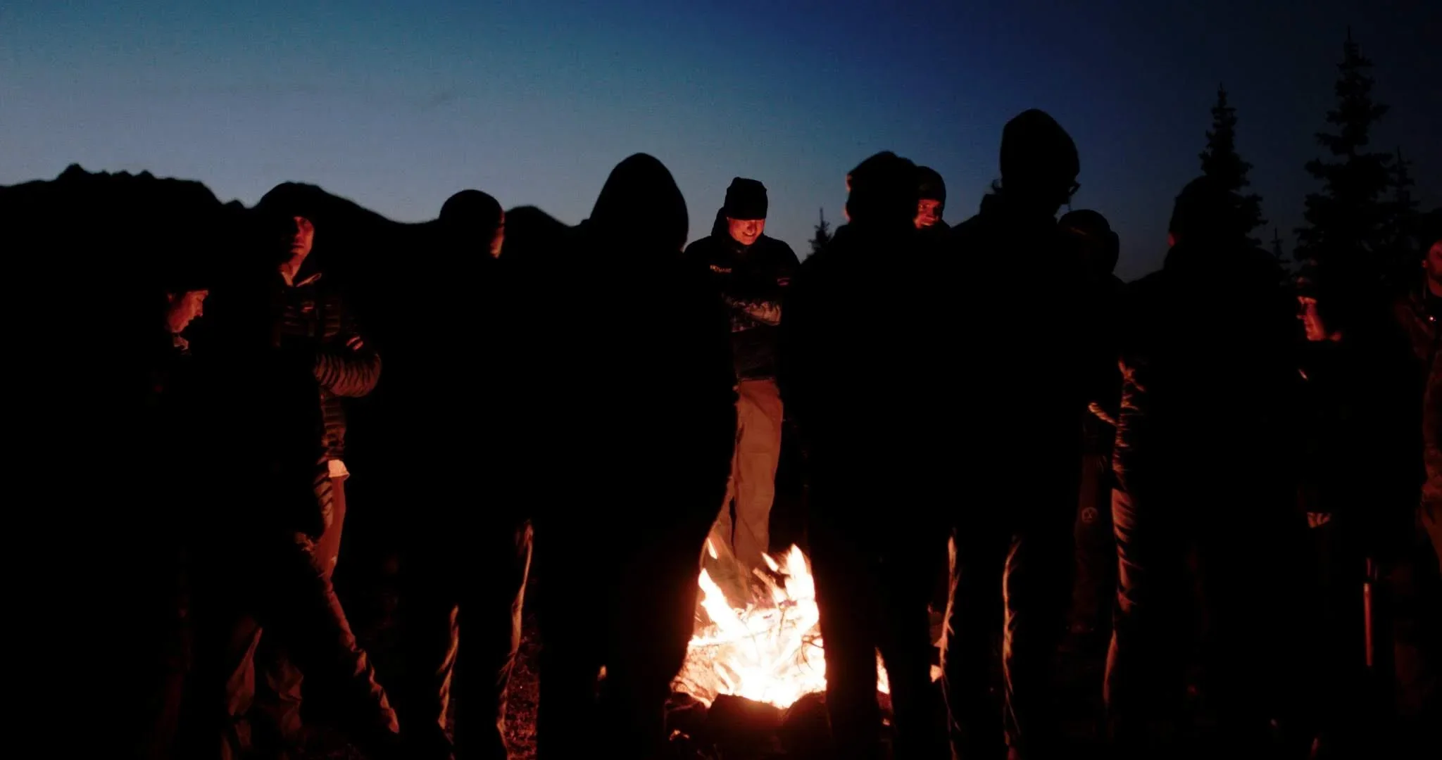 A group of people gathered around a campfire at night outdoors, with mountains and trees in the background.