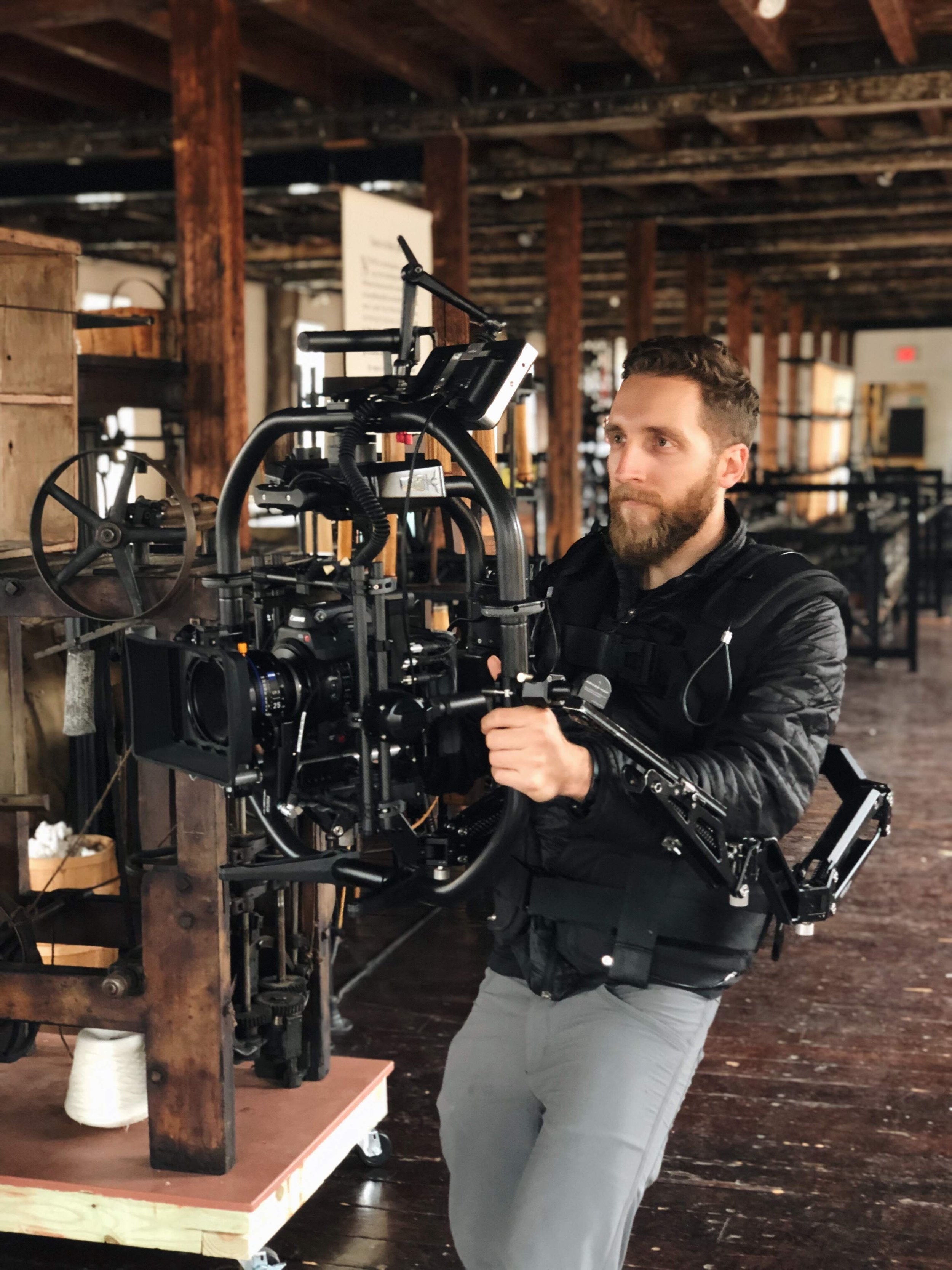 A man operating a camera rig on a film set, with wooden beams and shelves in the background.