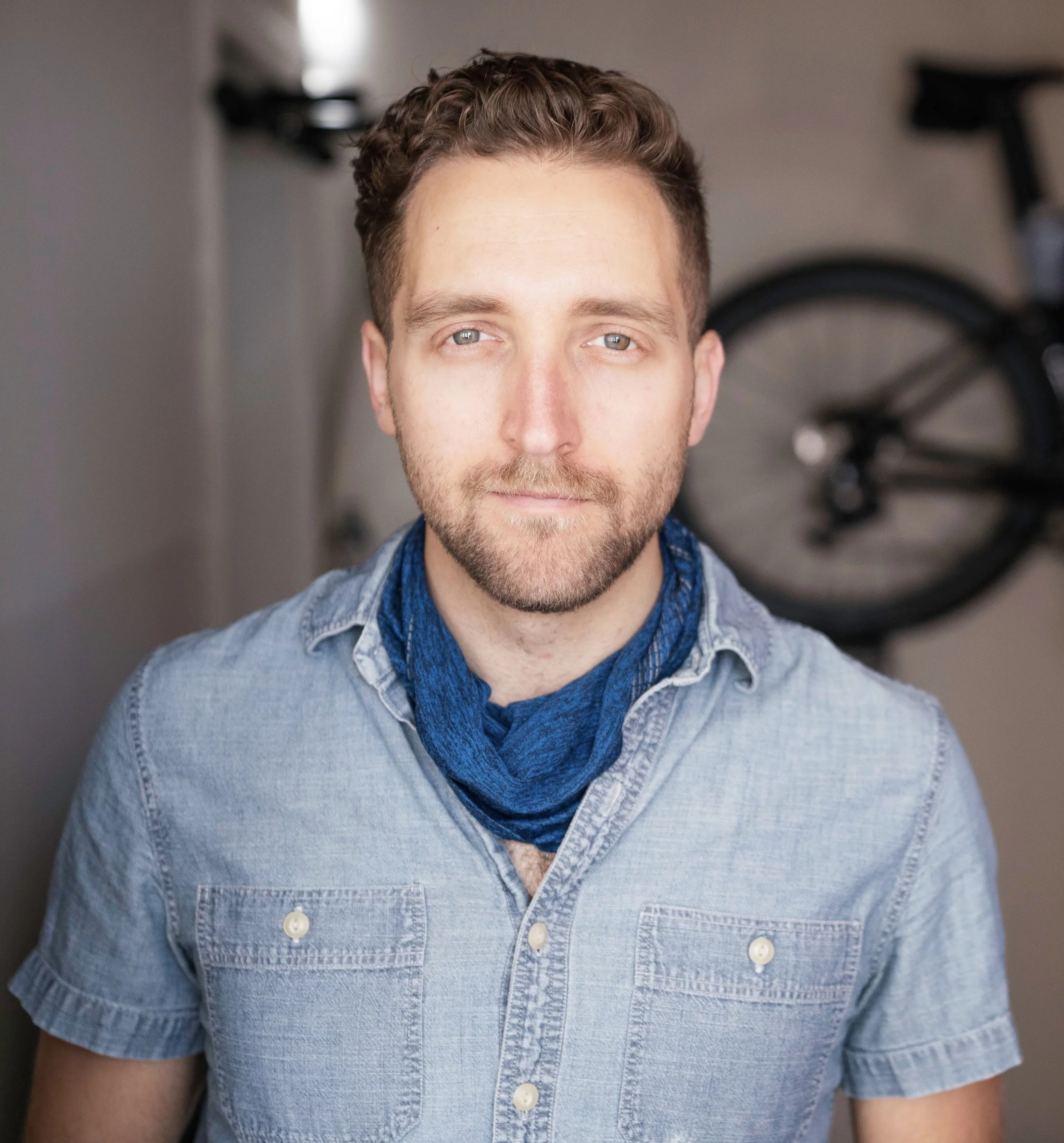 Close-up of a man with short, curly brown hair and light skin, wearing a denim shirt and a blue scarf, standing in front of a blurred background with a bicycle.