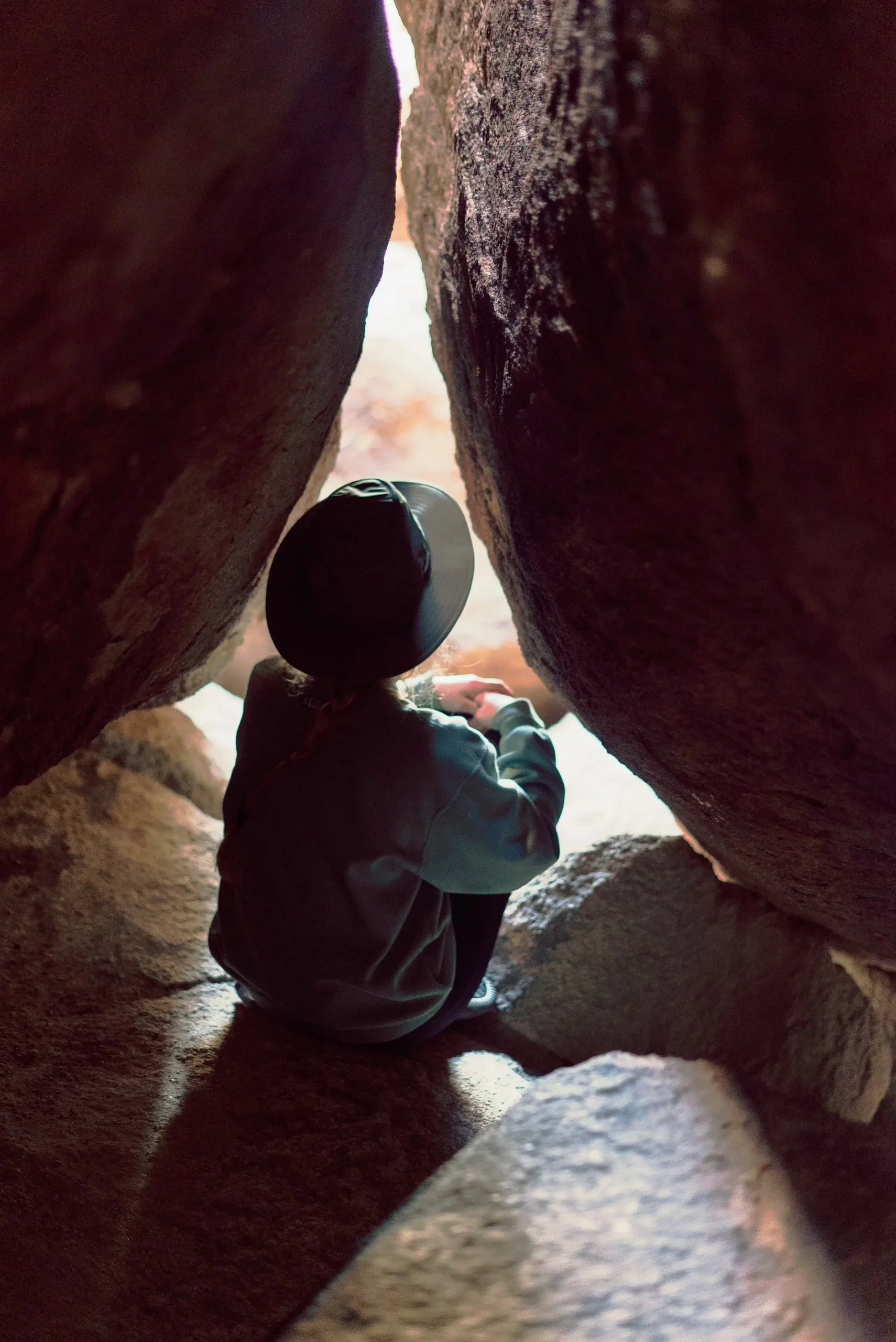 A person wearing a hat and jacket sitting between rocks, viewed from below, with light shining in from the outside.
