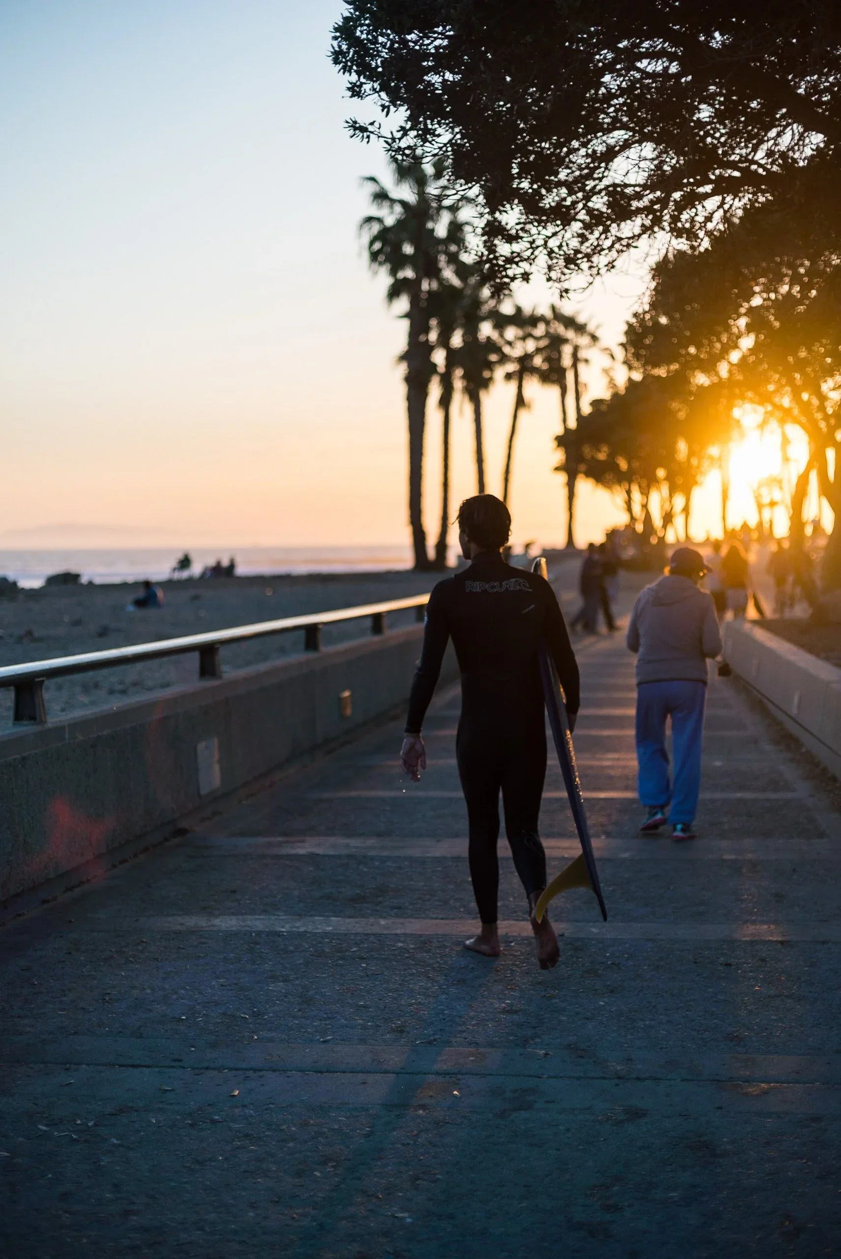 People walking along a beach promenade at sunset, with trees and the ocean in the background. One person is carrying a surfboard.