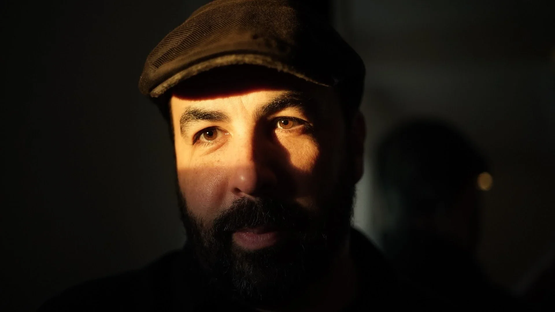 Close-up portrait of a man with a beard, wearing a cap, illuminated by warm light from one side, with a dark background and shadows on part of his face.