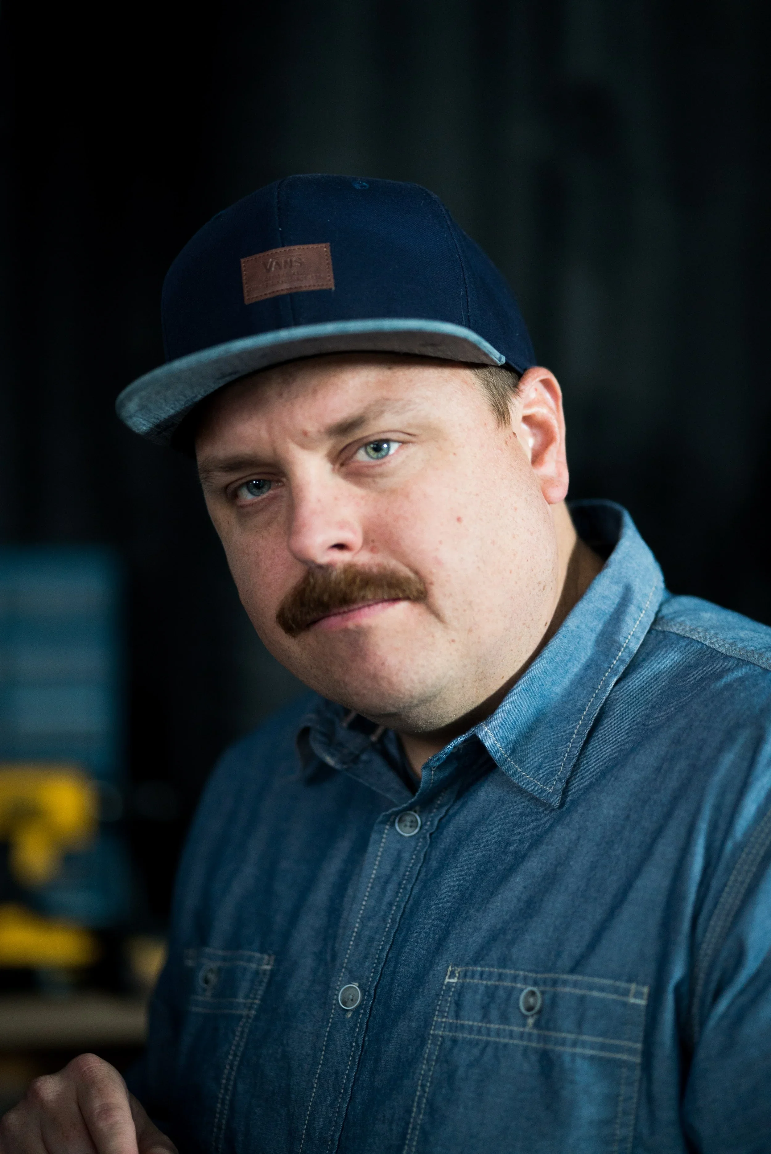 Close-up of a man with a mustache wearing a blue denim shirt and a navy blue cap with a brown leather patch. He has blue eyes and light skin, and is looking at the camera against a dark background.