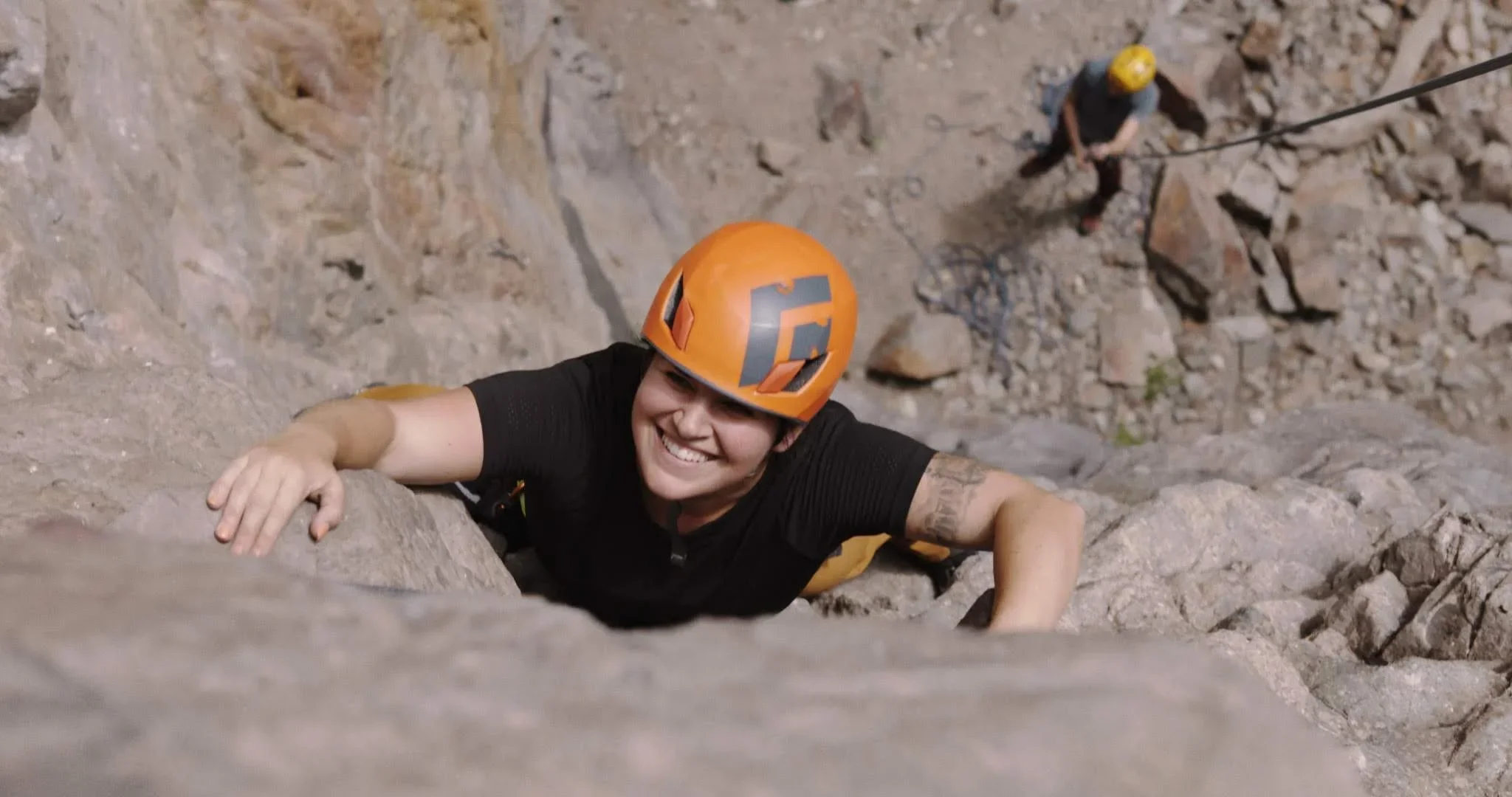 A woman wearing a safety helmet rock climbing on a steep rocky surface with a smiling face, with a person in the background securing the rope.