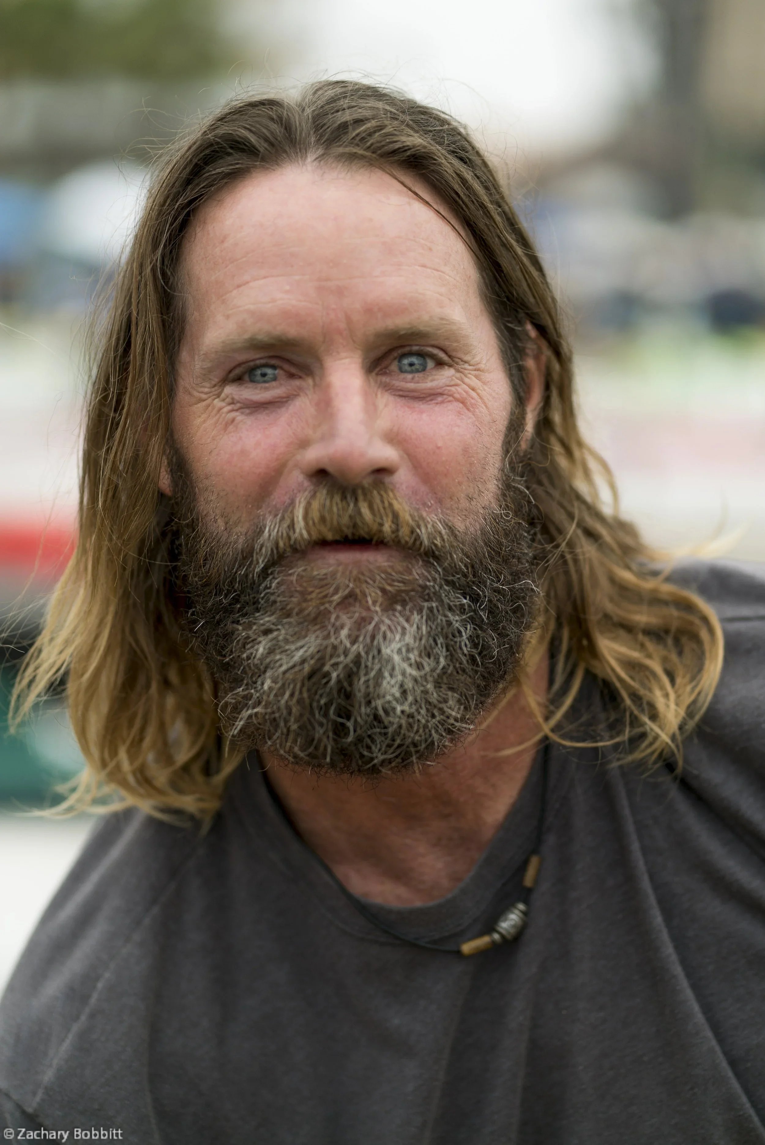 Close-up of a man with long hair and a beard, wearing a dark shirt and a necklace with beads, outdoors during daytime.
