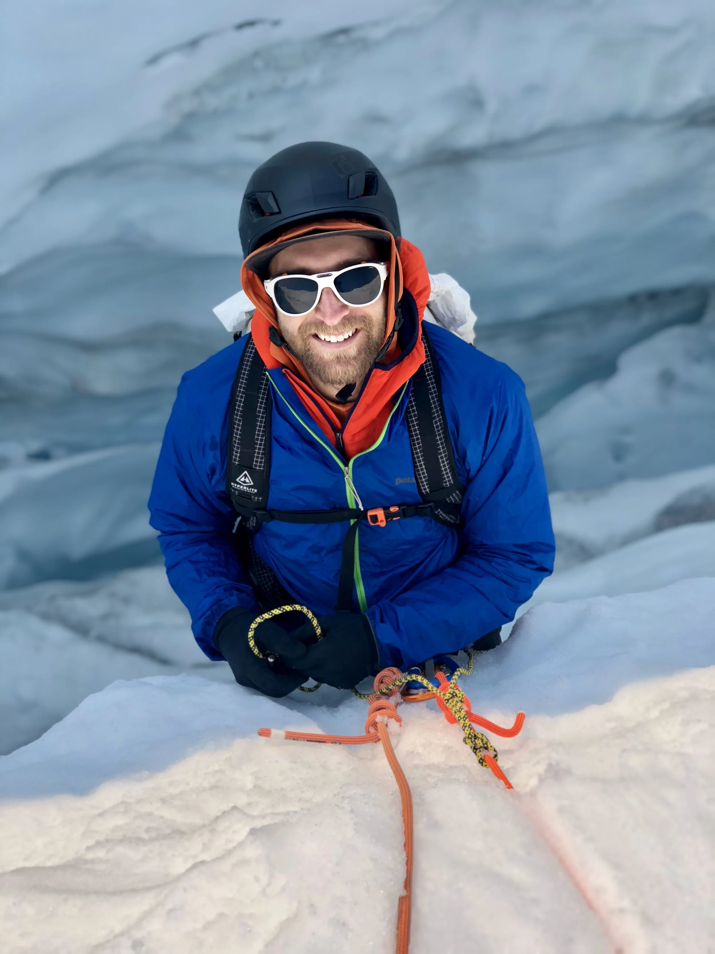 A man with a beard wearing sunglasses, an orange hoodie, a blue jacket, and a black helmet, smiling while sitting on snow in a snowy mountain landscape, holding an orange climbing rope.