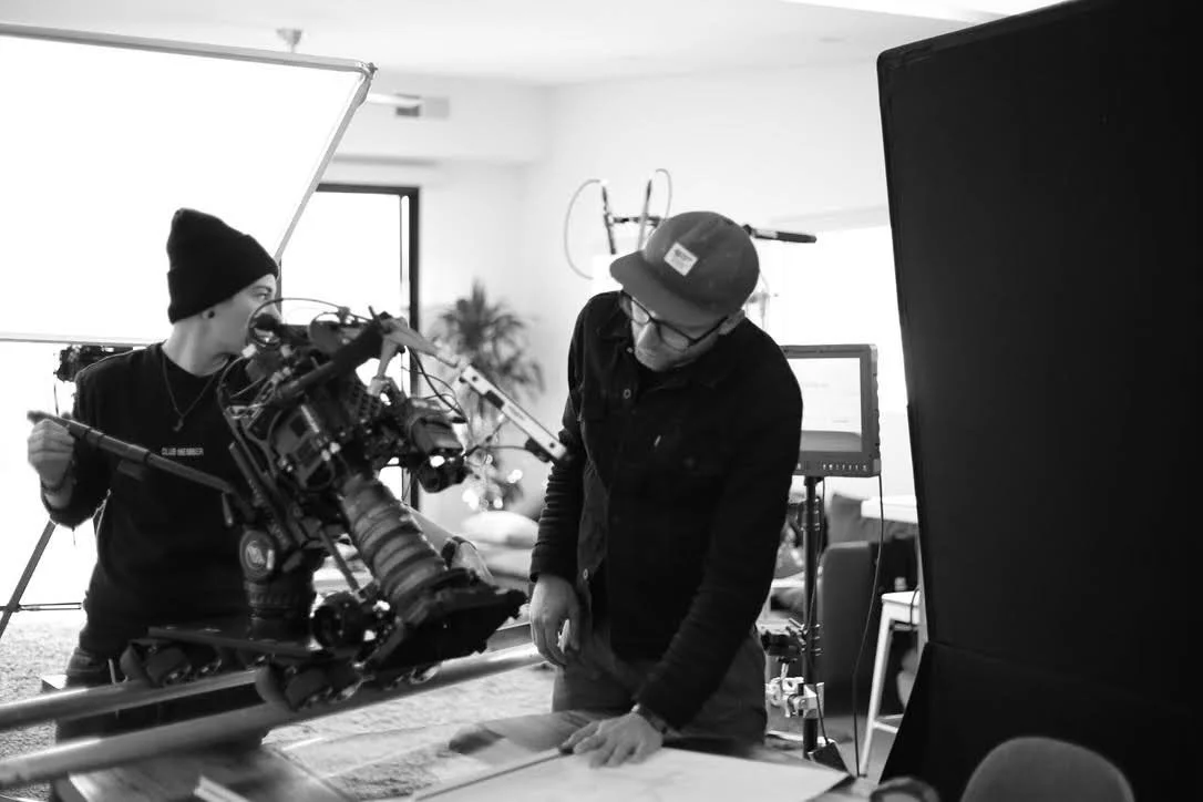 Two filmmakers on a set, one adjusting a camera on a dolly, the other looking down at a table.