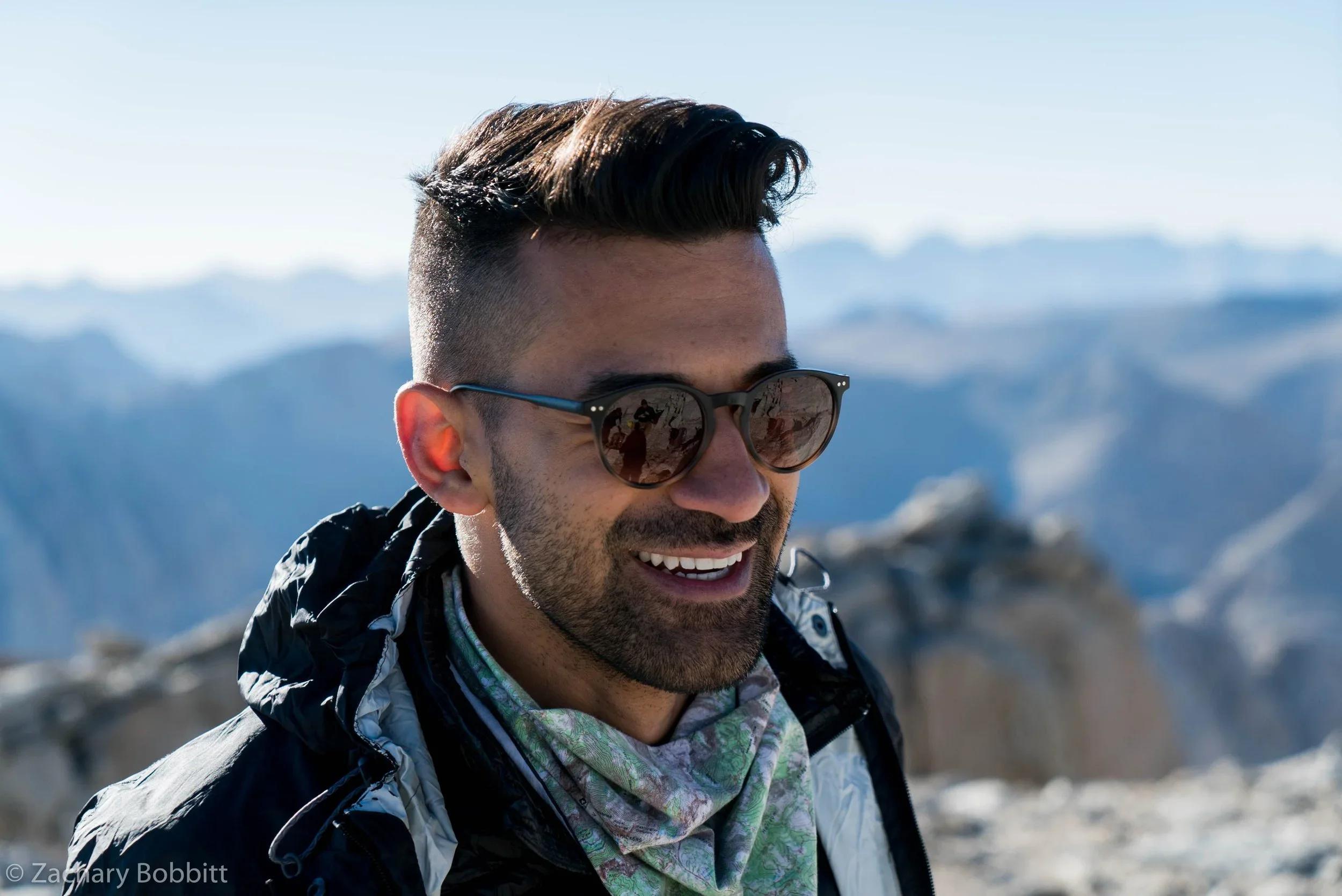 A smiling man wearing sunglasses, a patterned bandana, and outdoor clothing, standing in a snowy mountain landscape with blue sky and distant mountain peaks.
