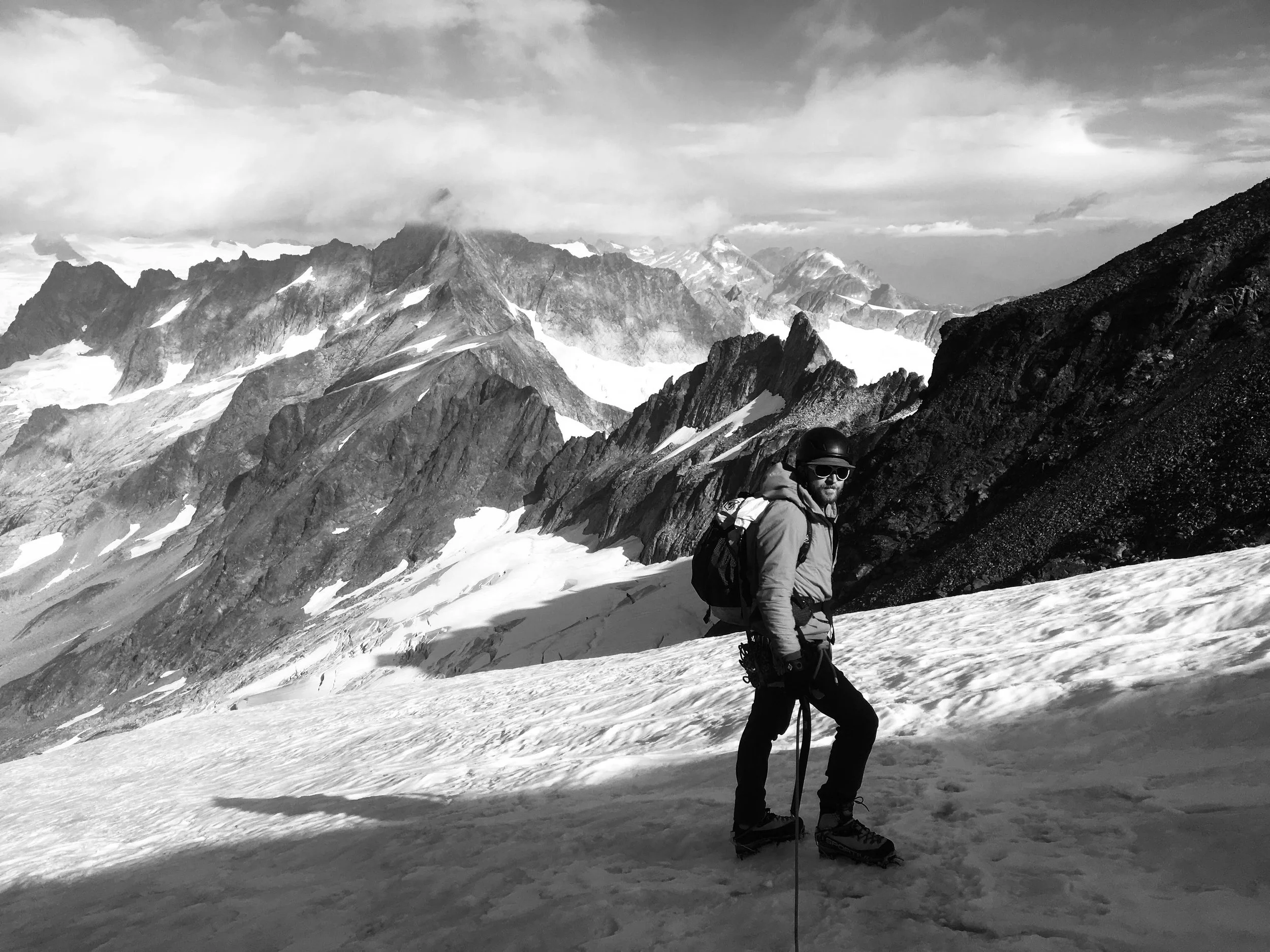 A person wearing a jacket, helmet, and sunglasses standing on snow-covered terrain with mountain peaks in the background.