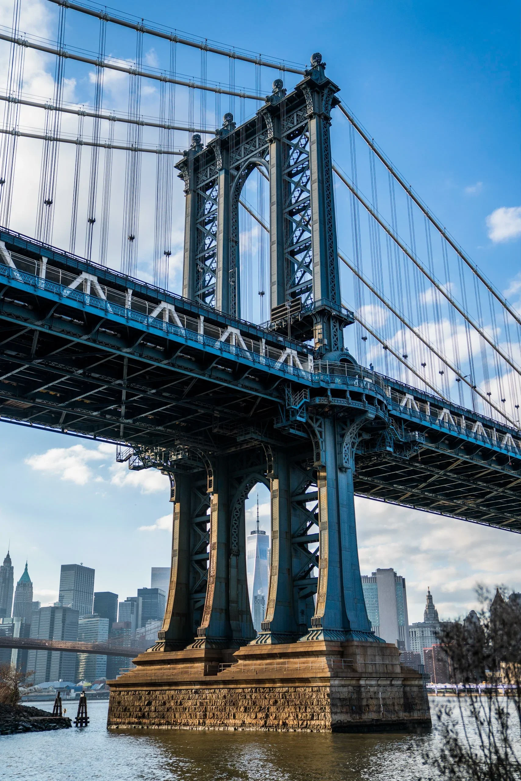 A view of the Manhattan Bridge in New York City with the city skyline in the background, including One World Trade Center, under a partly cloudy sky.