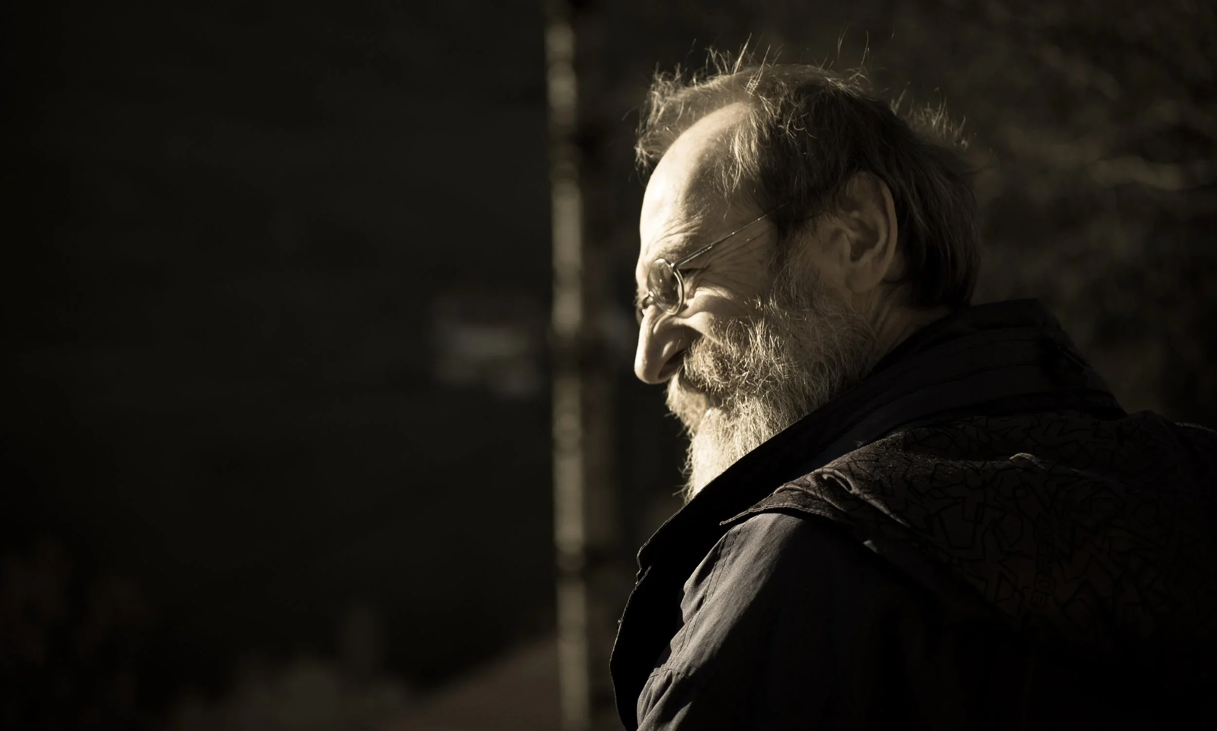 An elderly man with glasses and a beard, smiling and looking downward, outdoors in sunlight.