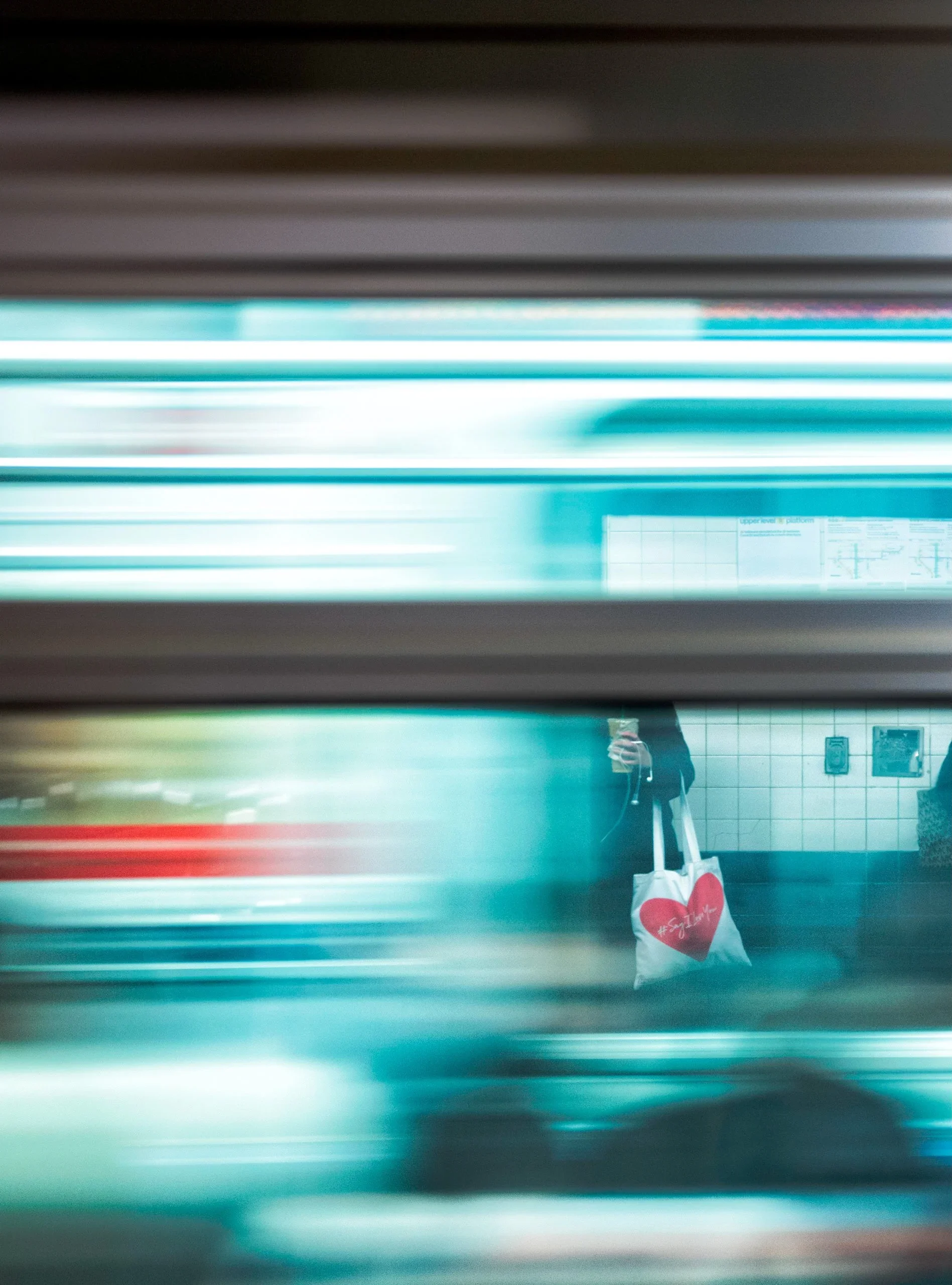 A person is taking a photo of themselves with a smartphone in front of a mirror in a subway station, while holding a tote bag with a red heart and the word 'Eat' on it.