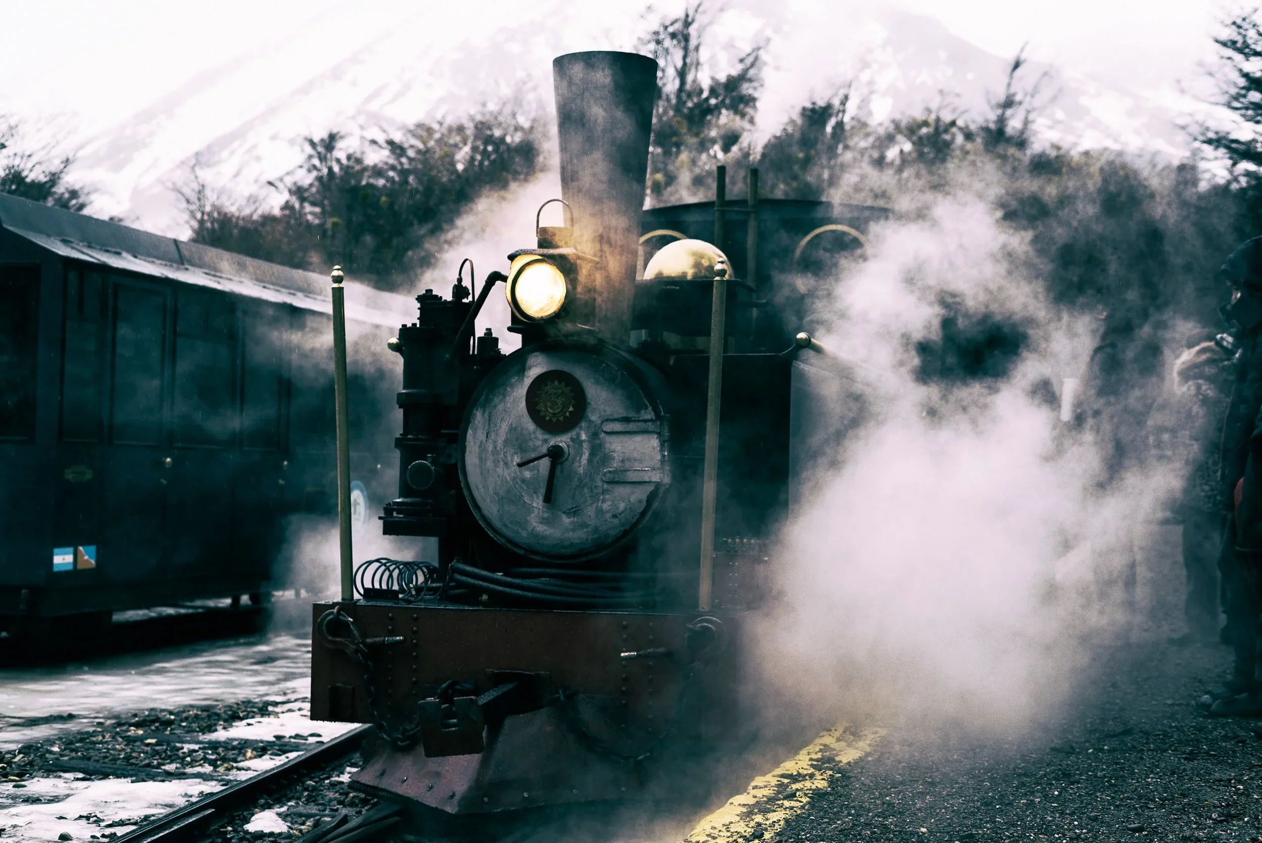 An old steam locomotive emitting steam as it moves along train tracks with a mountain and trees in the background.
