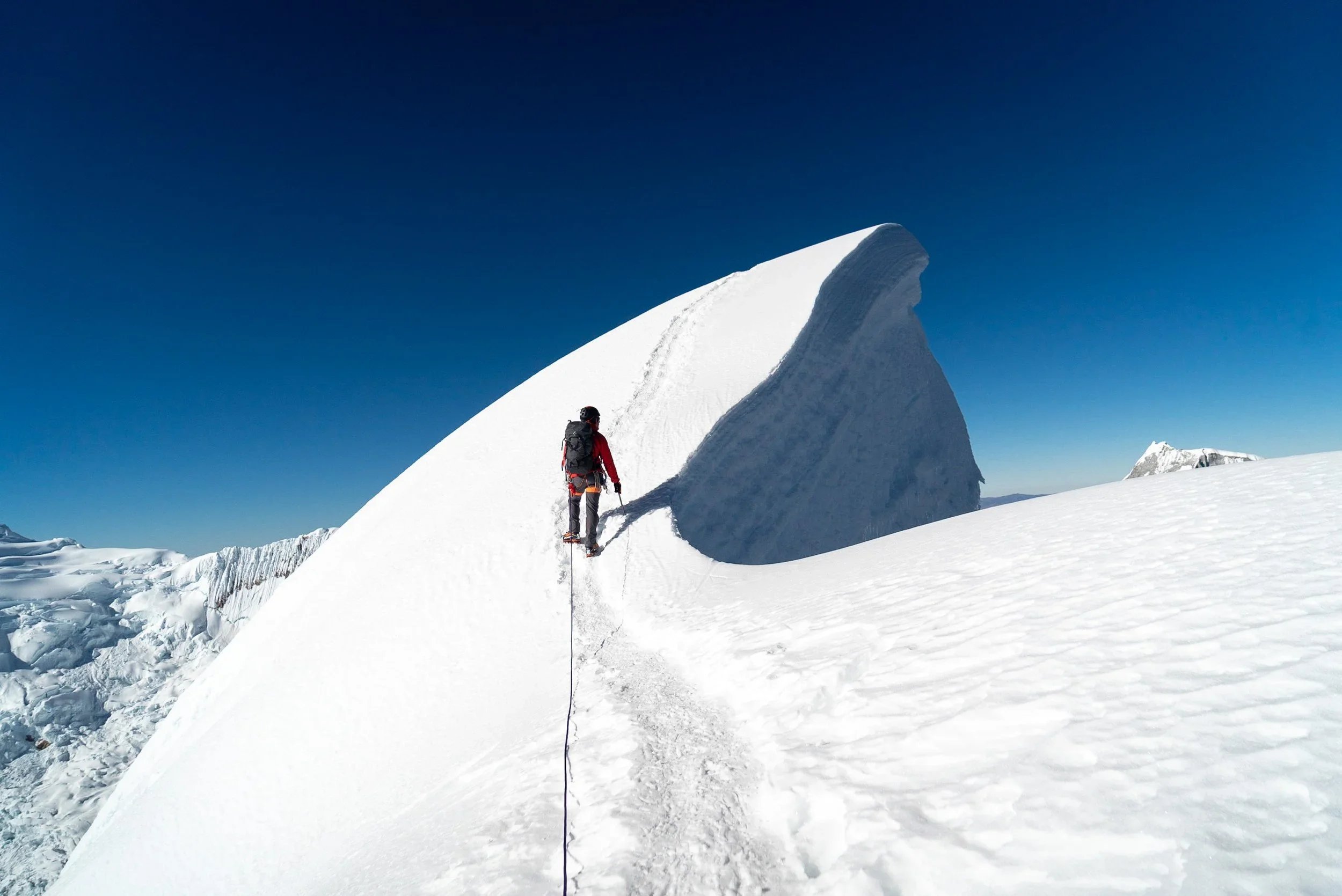 A person dressed in winter gear and carrying a backpack is climbing a snow-covered mountain ridge with a large snow formation and blue sky in the background.